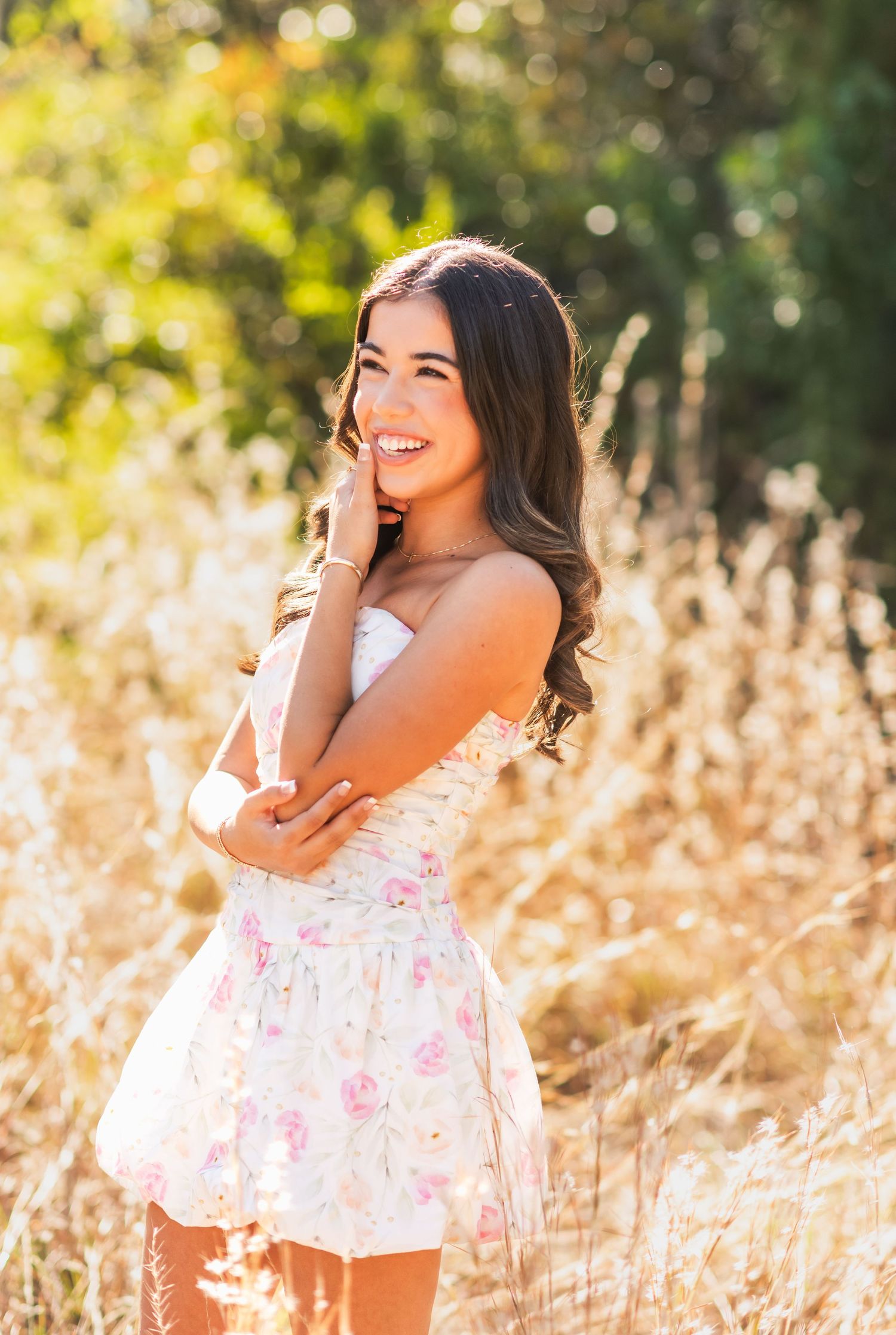 Young woman in floral sundress smiling in golden field, long brunette hair, outdoor senior portrait photography.