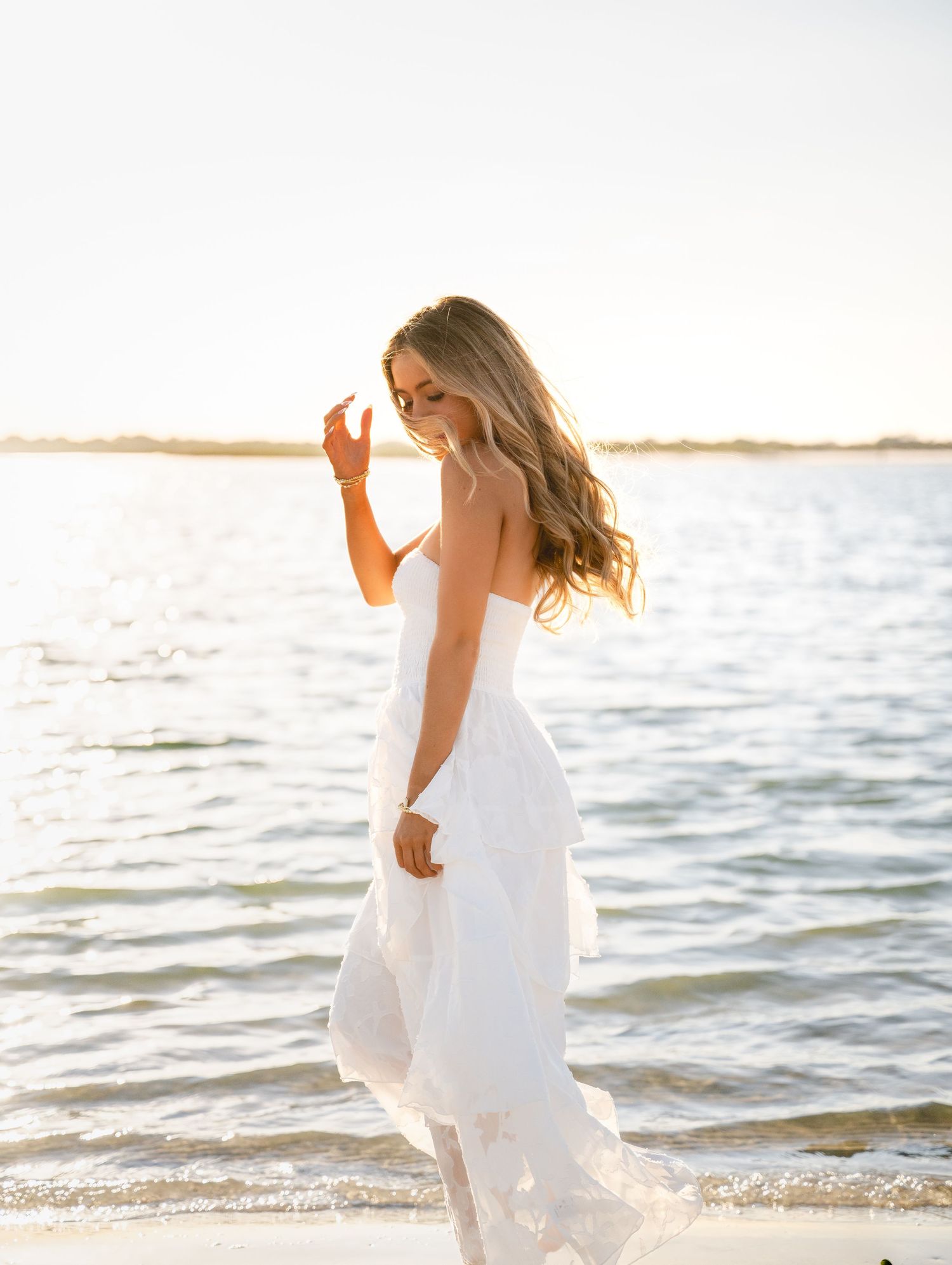 A woman in a flowing white dress walks along a sunlit beach at golden hour with gentle waves in the background.