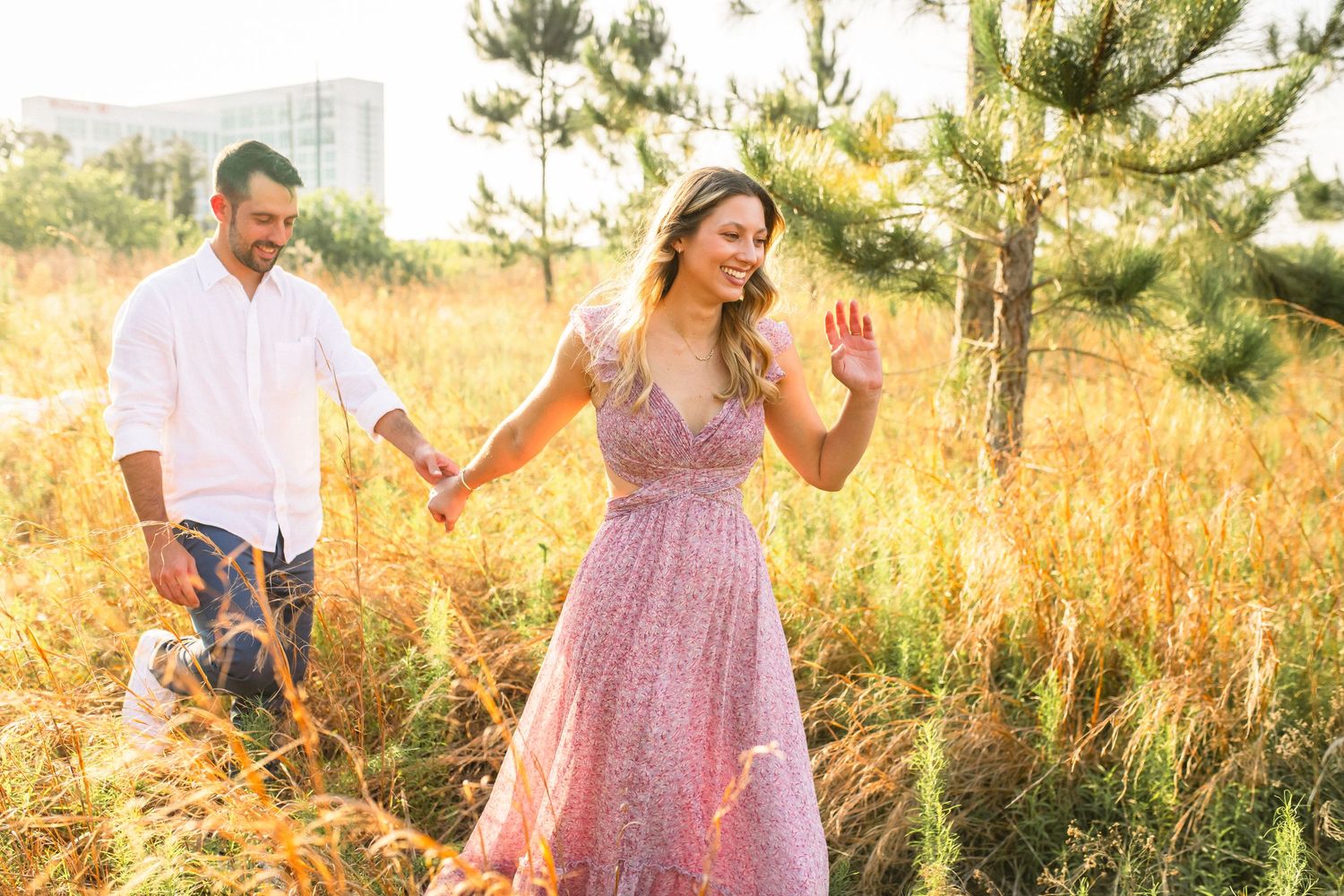 Two people walk through a golden field at sunset wearing summer attire, with pine trees in the background.