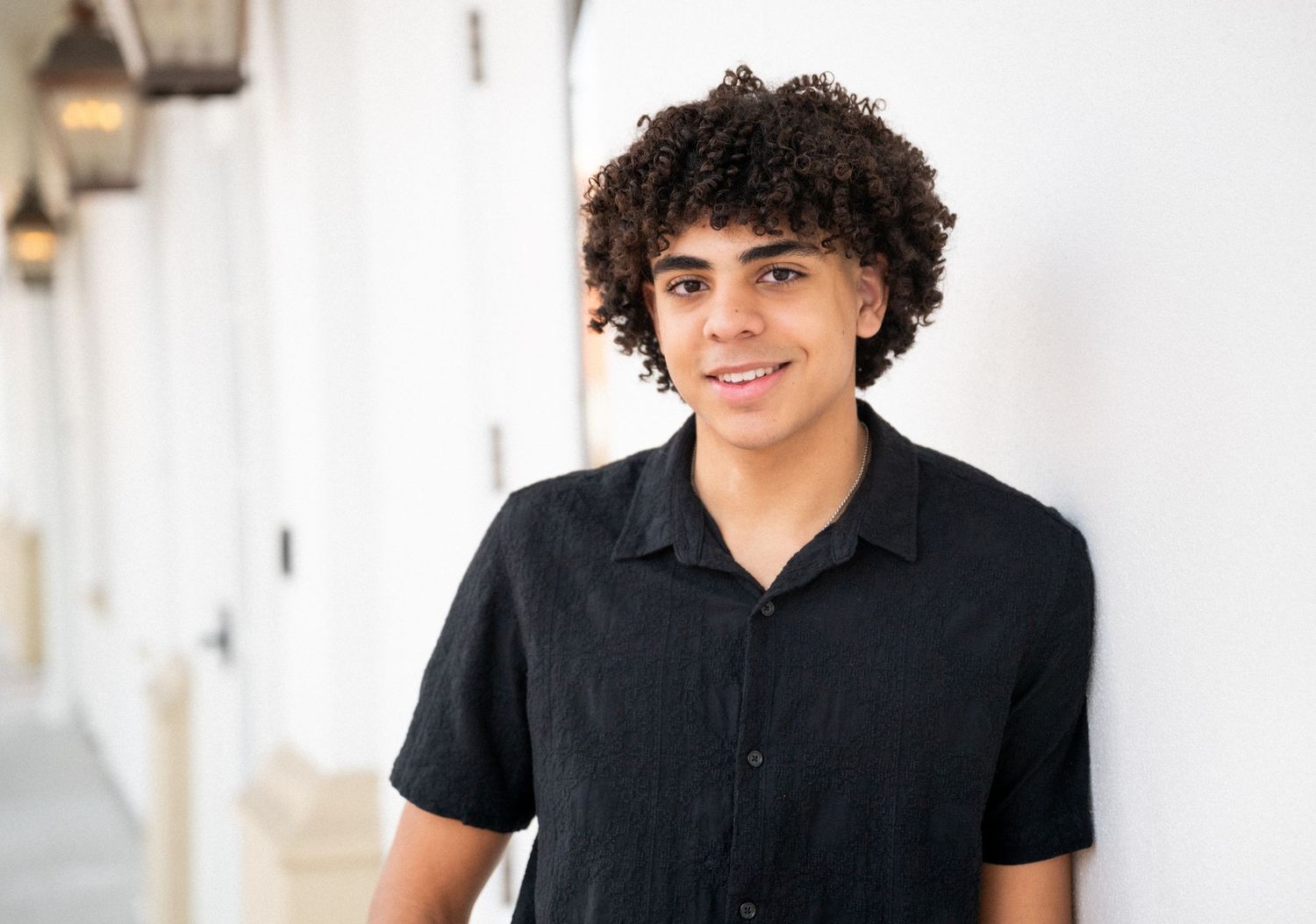 Smiling young man with curly hair wearing a black shirt, leaning against a white wall outdoors.