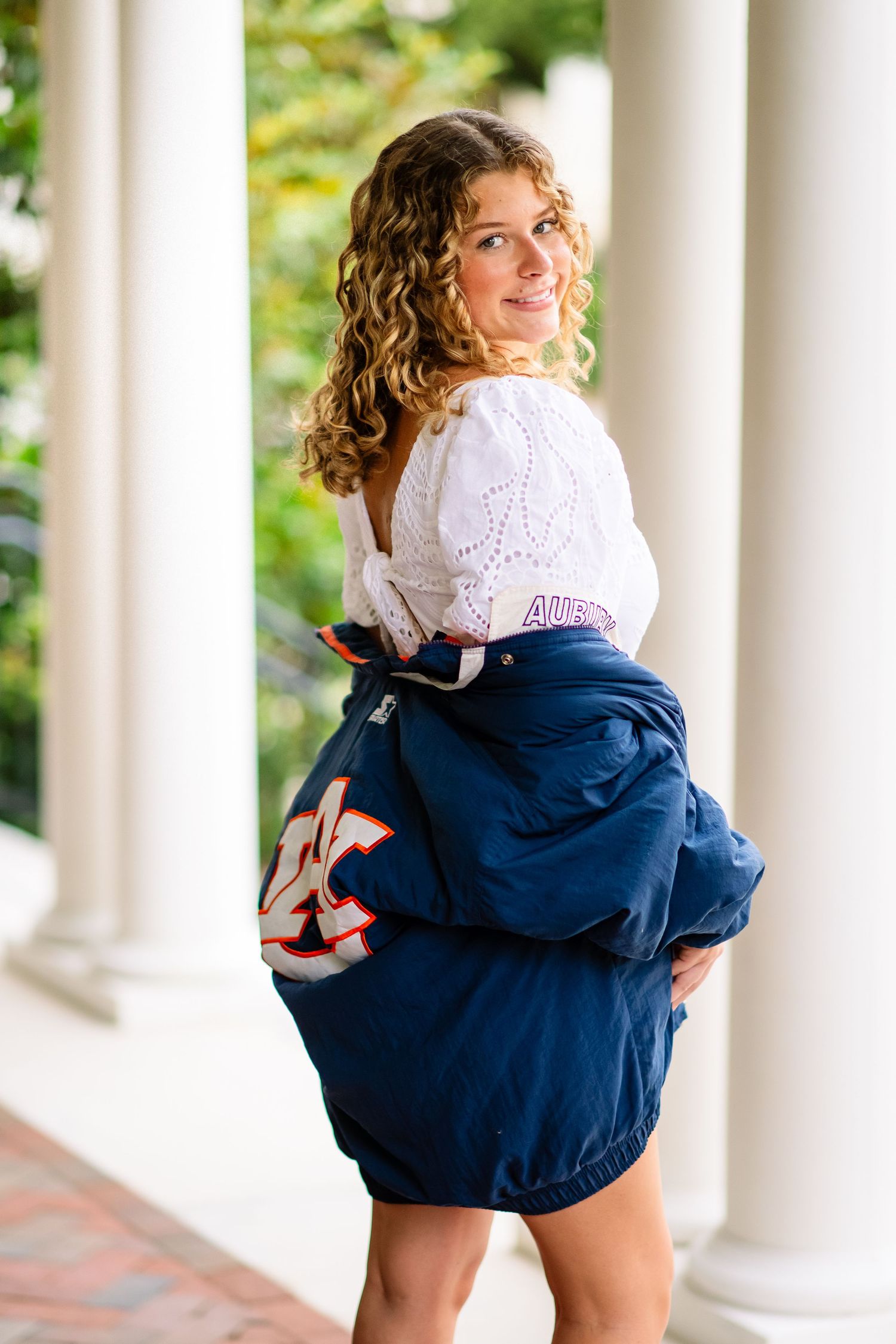 Young woman with curly hair posing in a white top and navy Auburn University jacket between white columns.