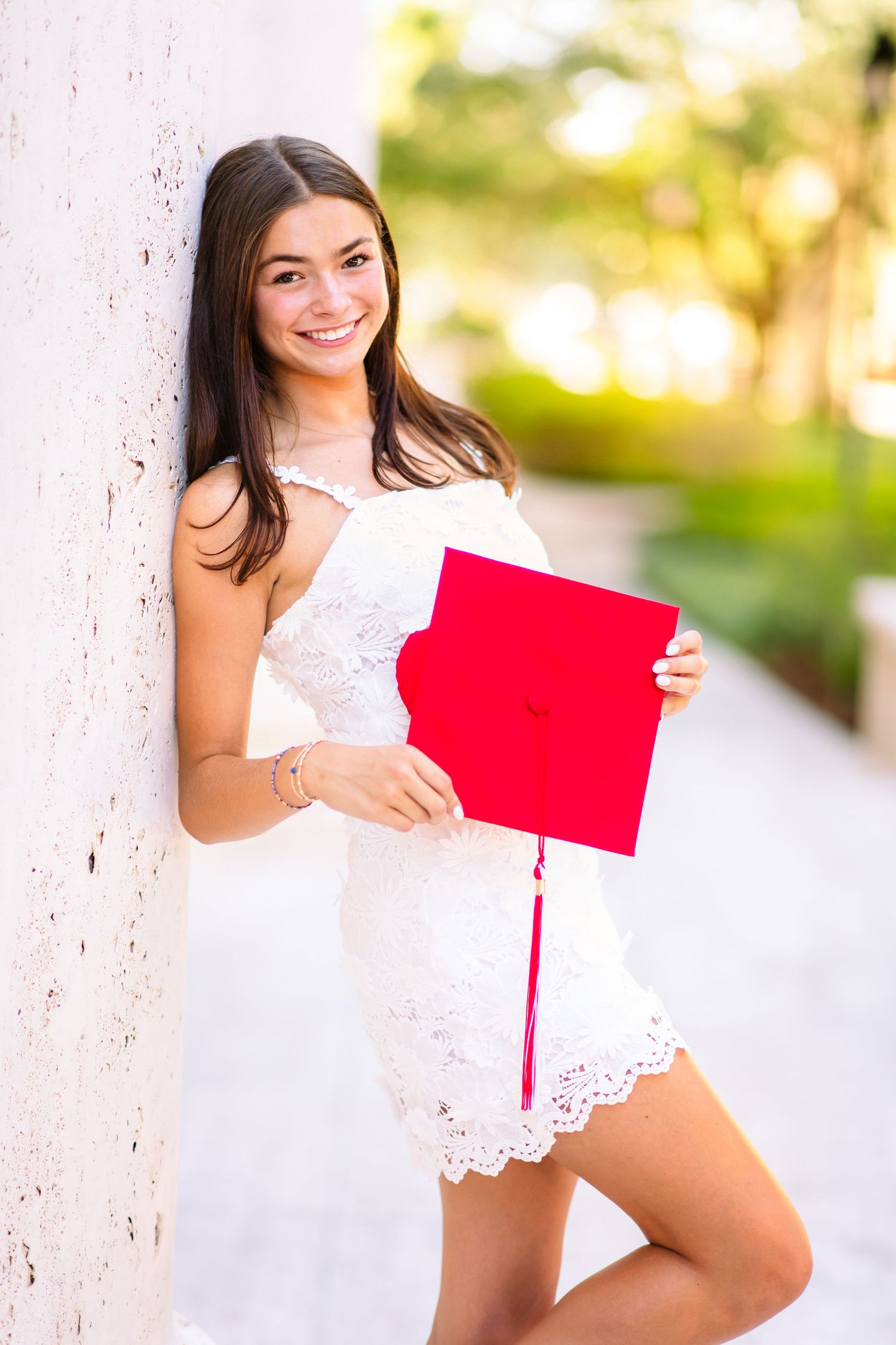 Smiling young woman in white lace dress holding red graduation cap, leaning against white column outdoors.