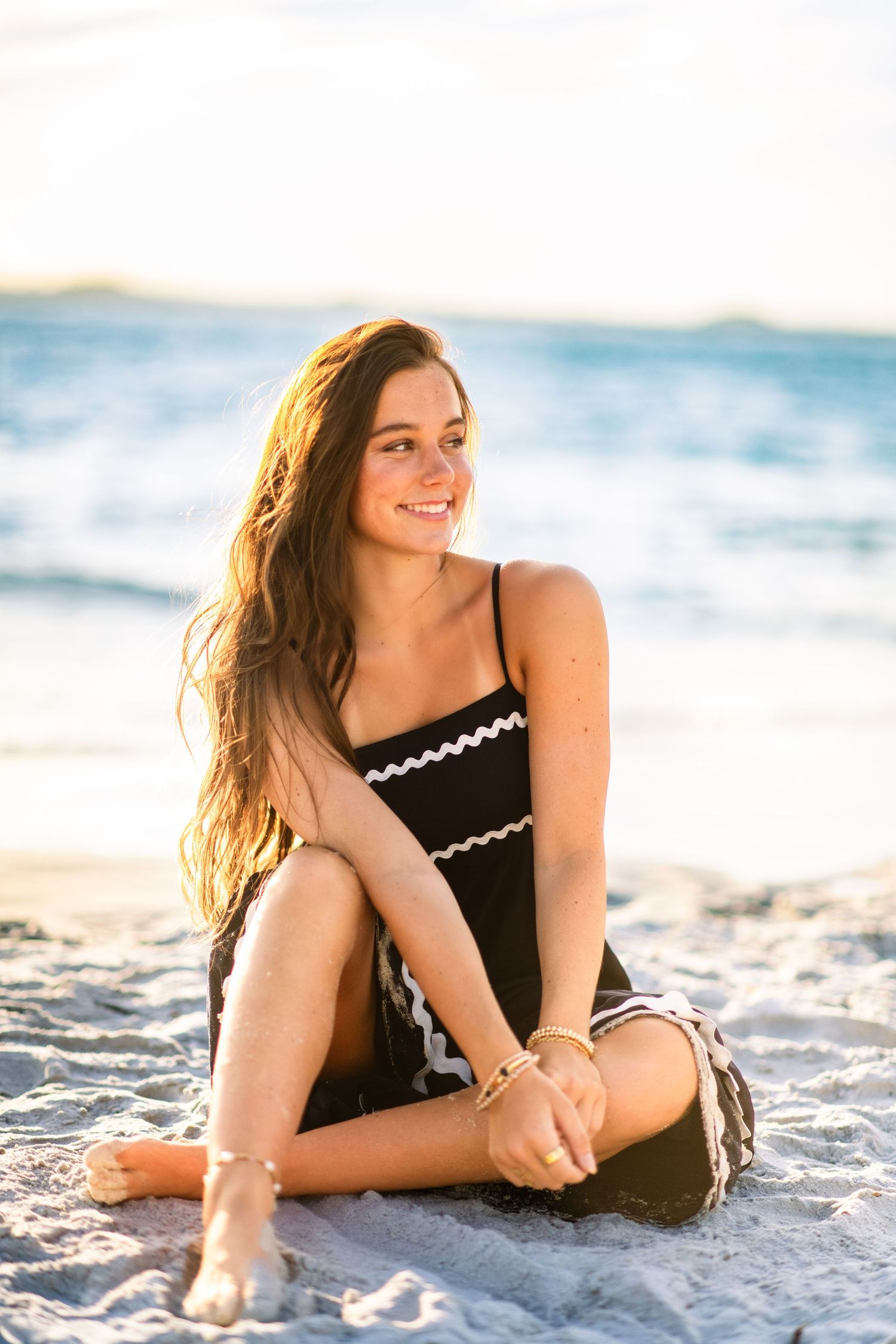 Young woman in black dress sitting on sandy beach smiling, with ocean waves in background during golden hour.