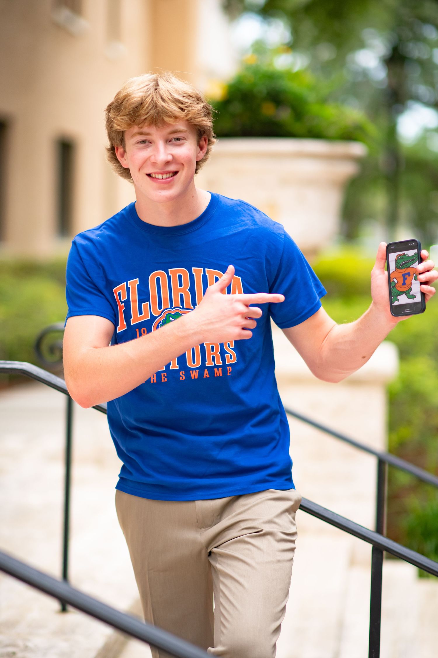 Smiling young man in Florida Gators blue t-shirt pointing at phone on outdoor steps.