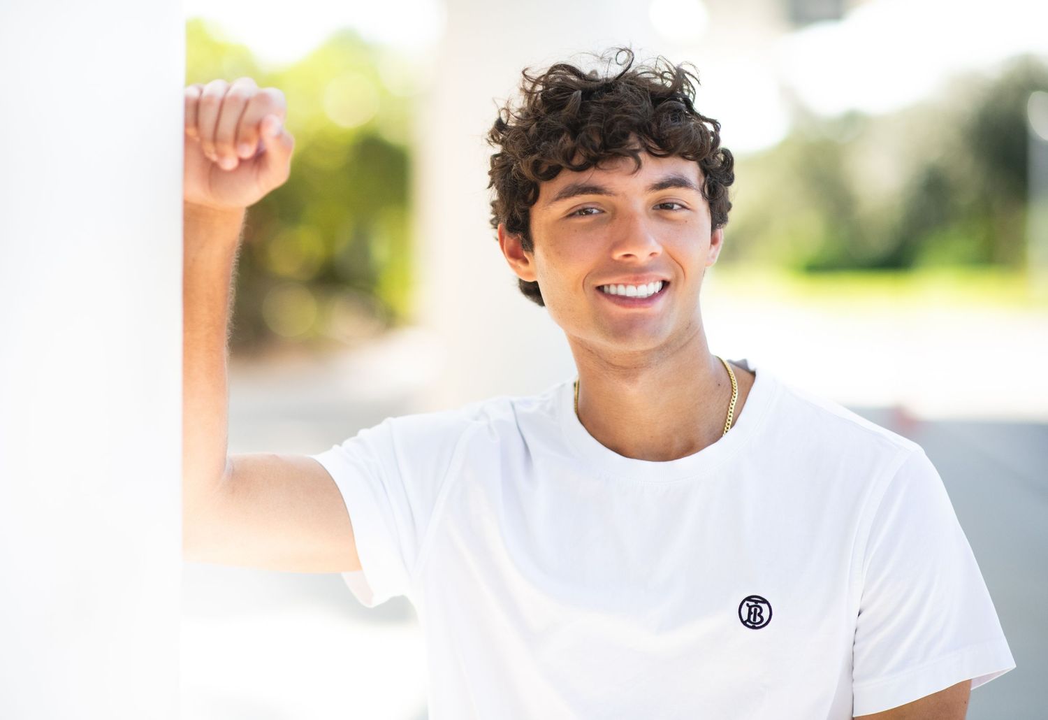 A person wearing a white t-shirt smiles while standing outdoors in bright natural lighting.