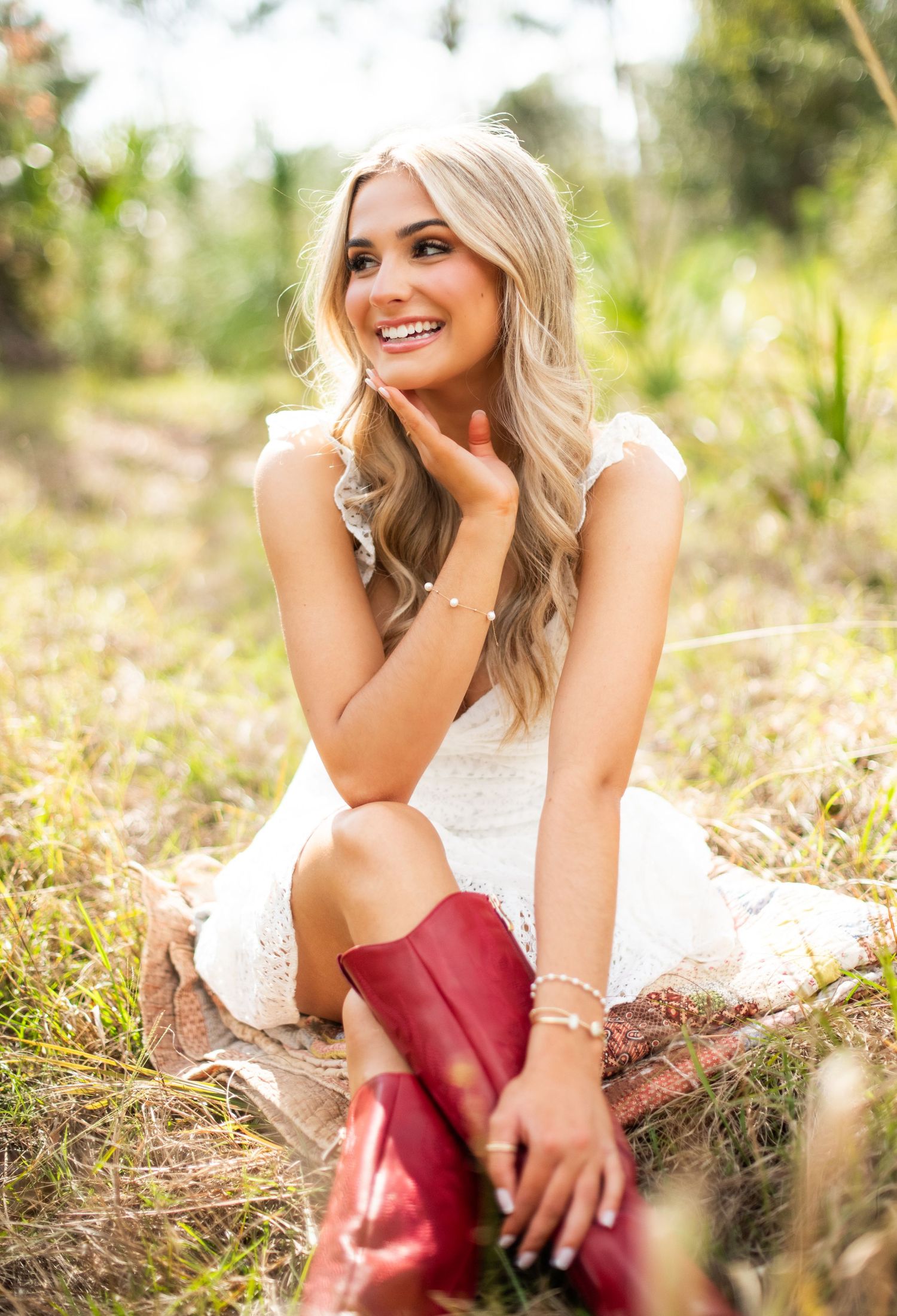 A person in a white summer dress sits in a field wearing red boots while posing outdoors.