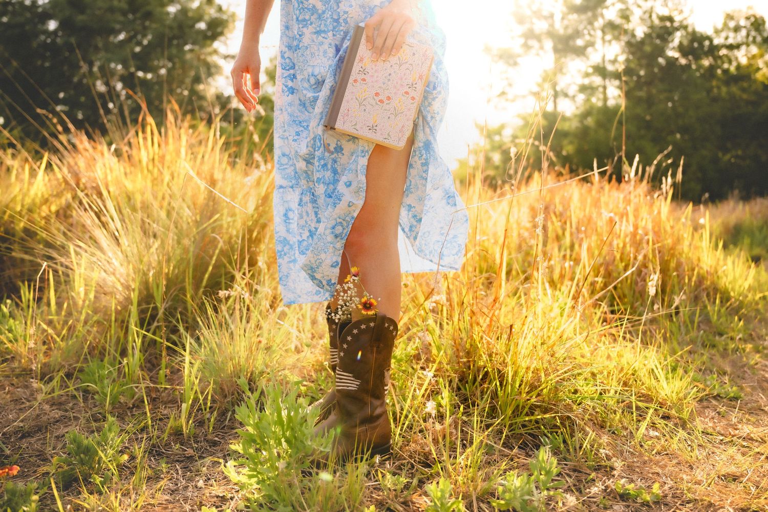 Woman in floral dress and brown boots standing in golden sunlit field holding a book.