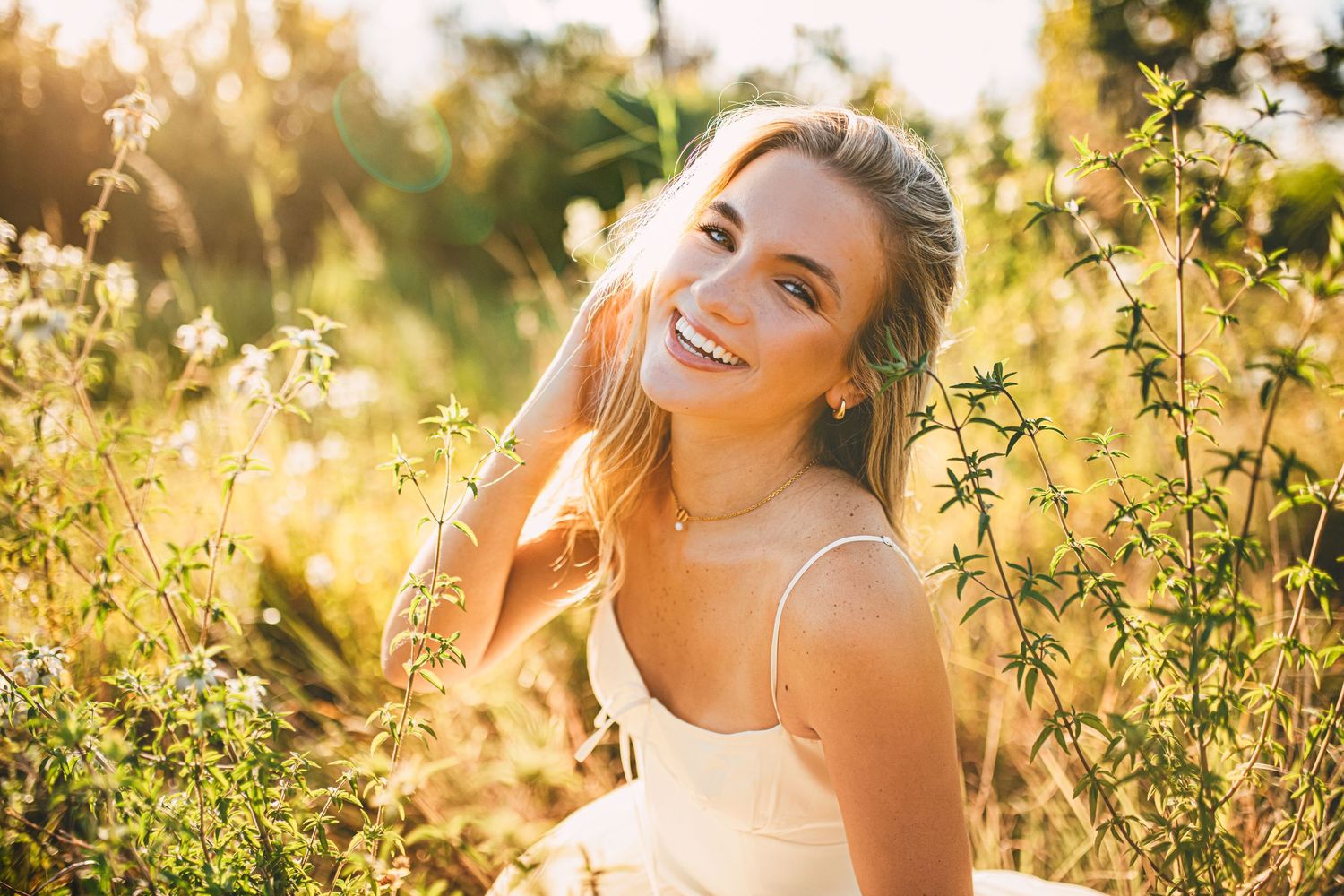 Young woman smiling in a sunlit field of wildflowers, wearing a white dress, golden hour light.