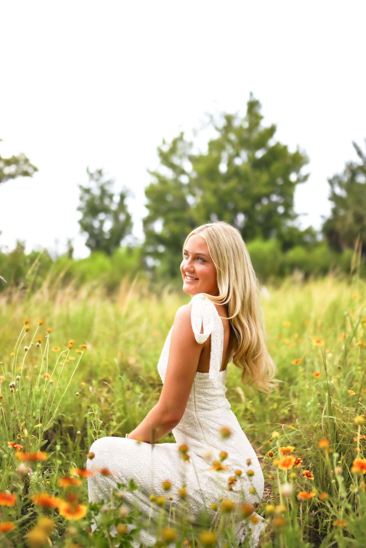 Smiling blonde woman in white dress sitting in a wildflower meadow with orange flowers and green trees.