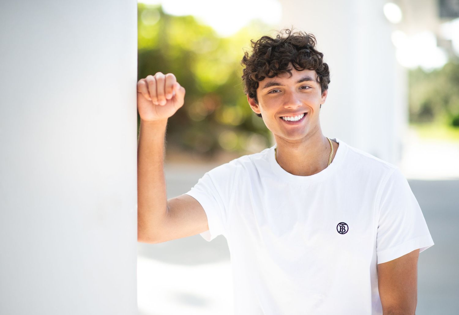 Smiling young man in white t-shirt leaning against a white pillar outdoors with blurred green background.