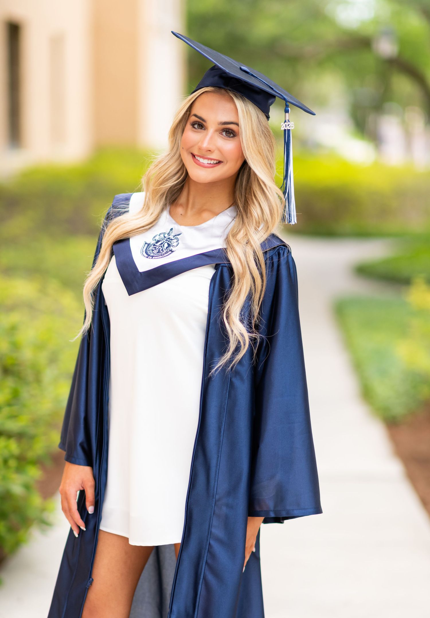Smiling blonde woman in navy blue graduation cap and gown poses outdoors on a sunny campus walkway.