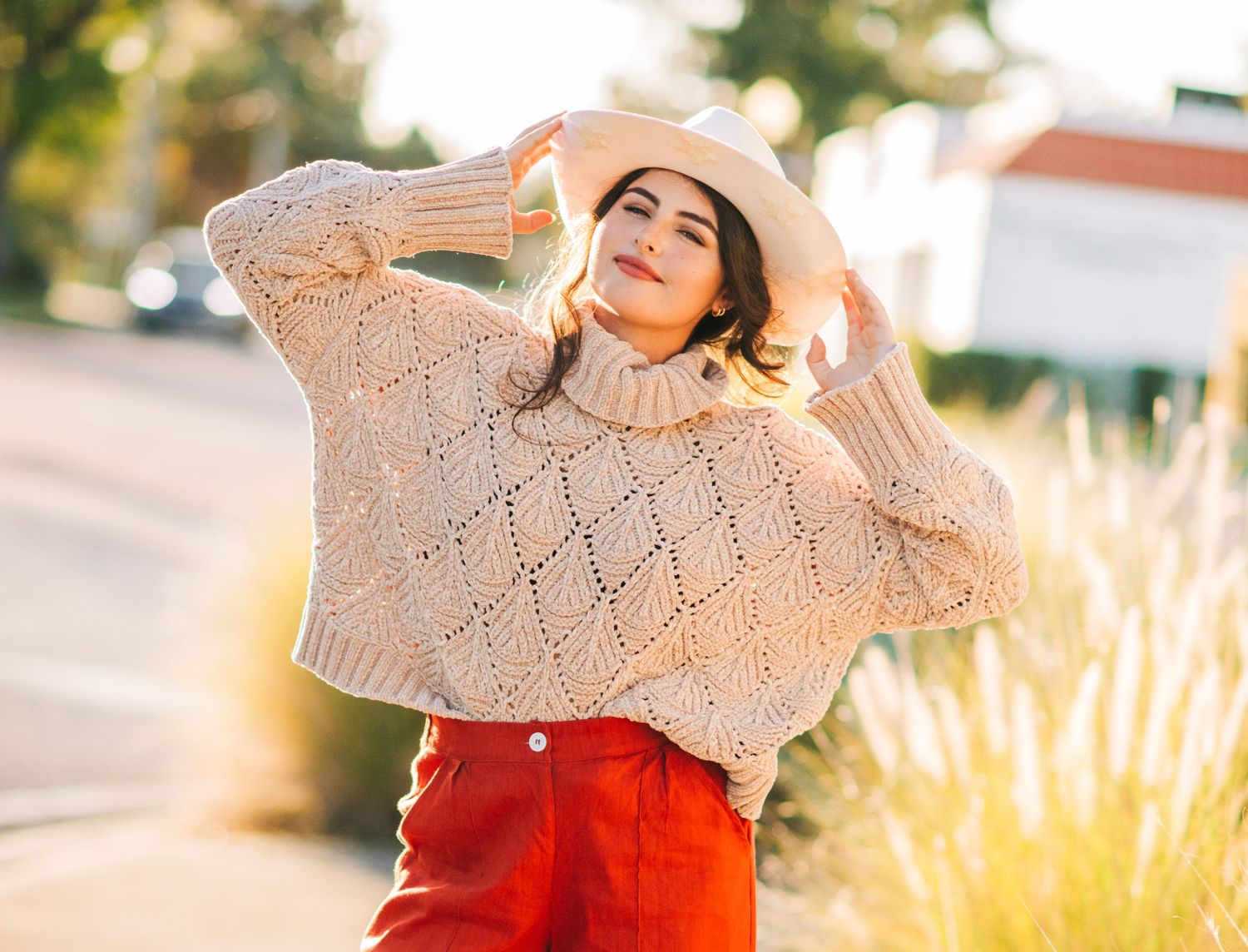 Trendy fashionista in beige sweater and red skirt poses with wide-brimmed hat against sunset backdrop.