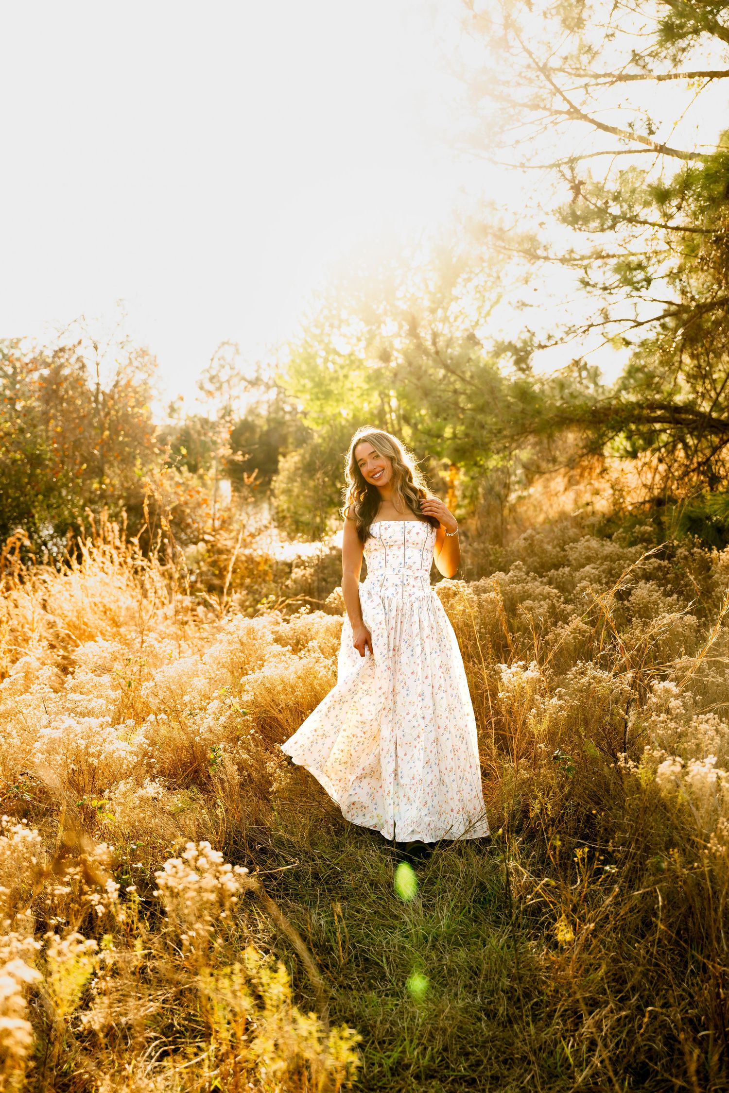 Young woman in white floral maxi dress standing in golden meadow with warm sunlight glowing through trees.