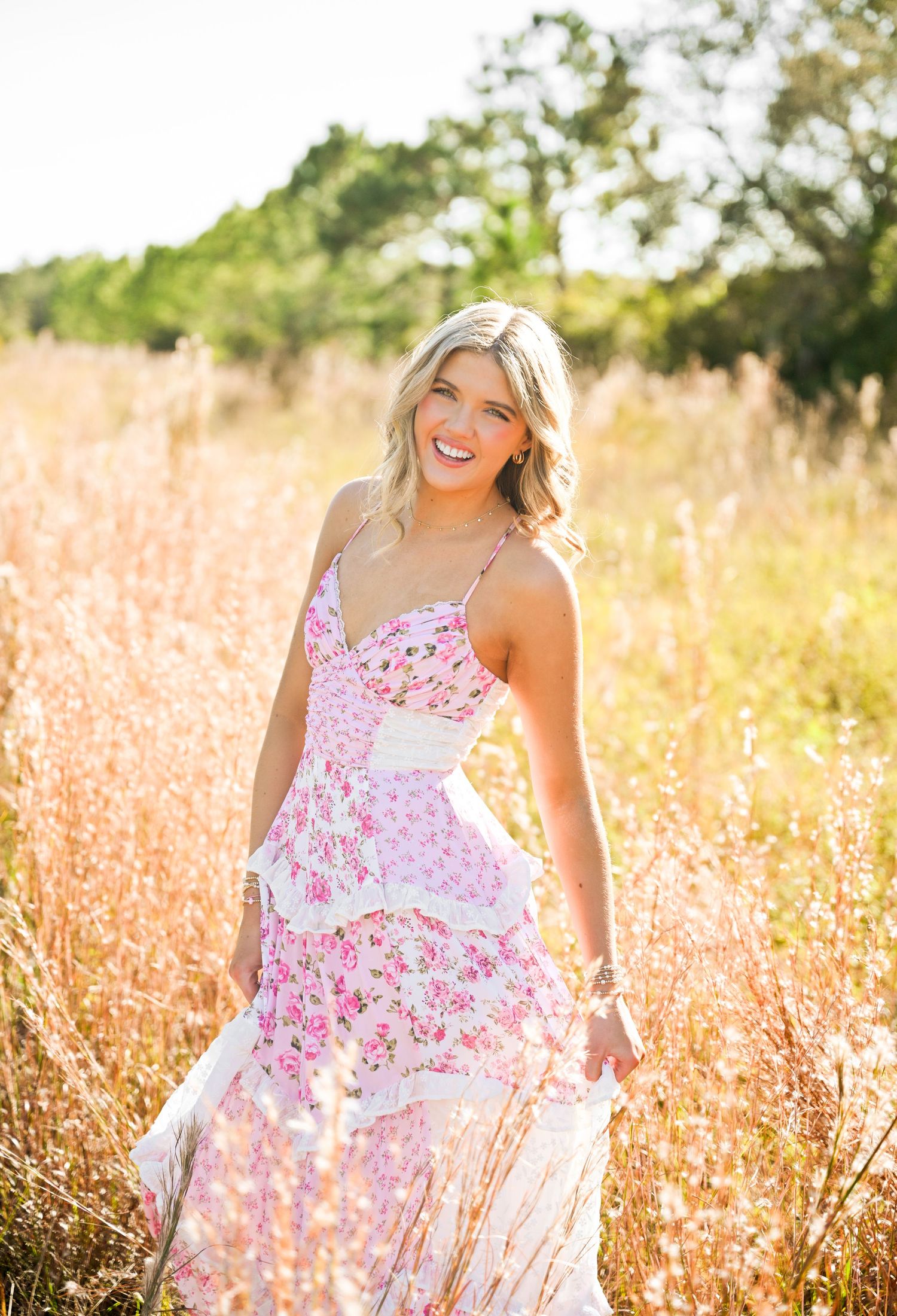 Smiling blonde woman in pink floral maxi dress standing in golden sunlit meadow with tall grass and trees.