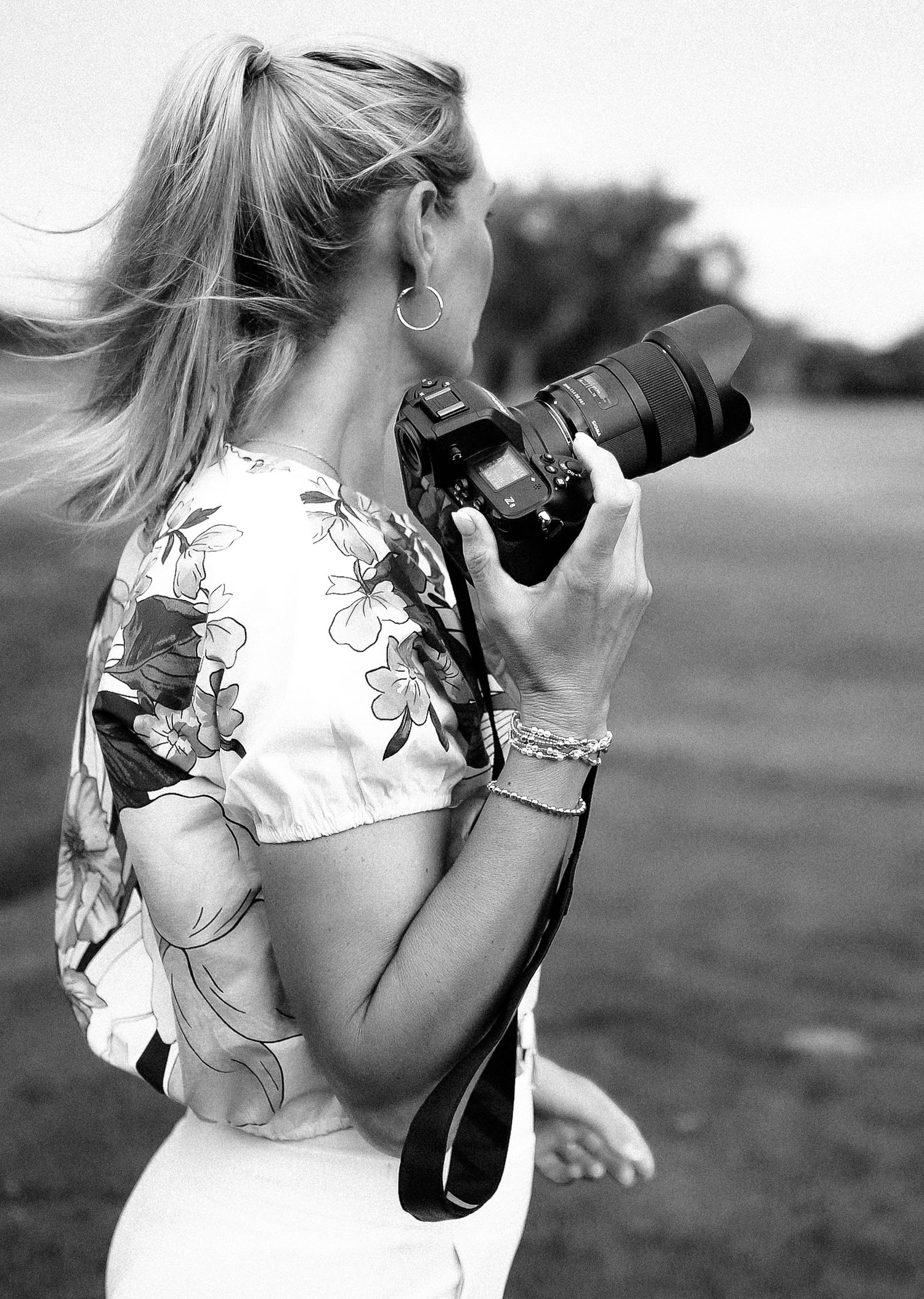 Black and white photo of a blonde woman photographing outdoors with a DSLR camera, wearing a floral top.