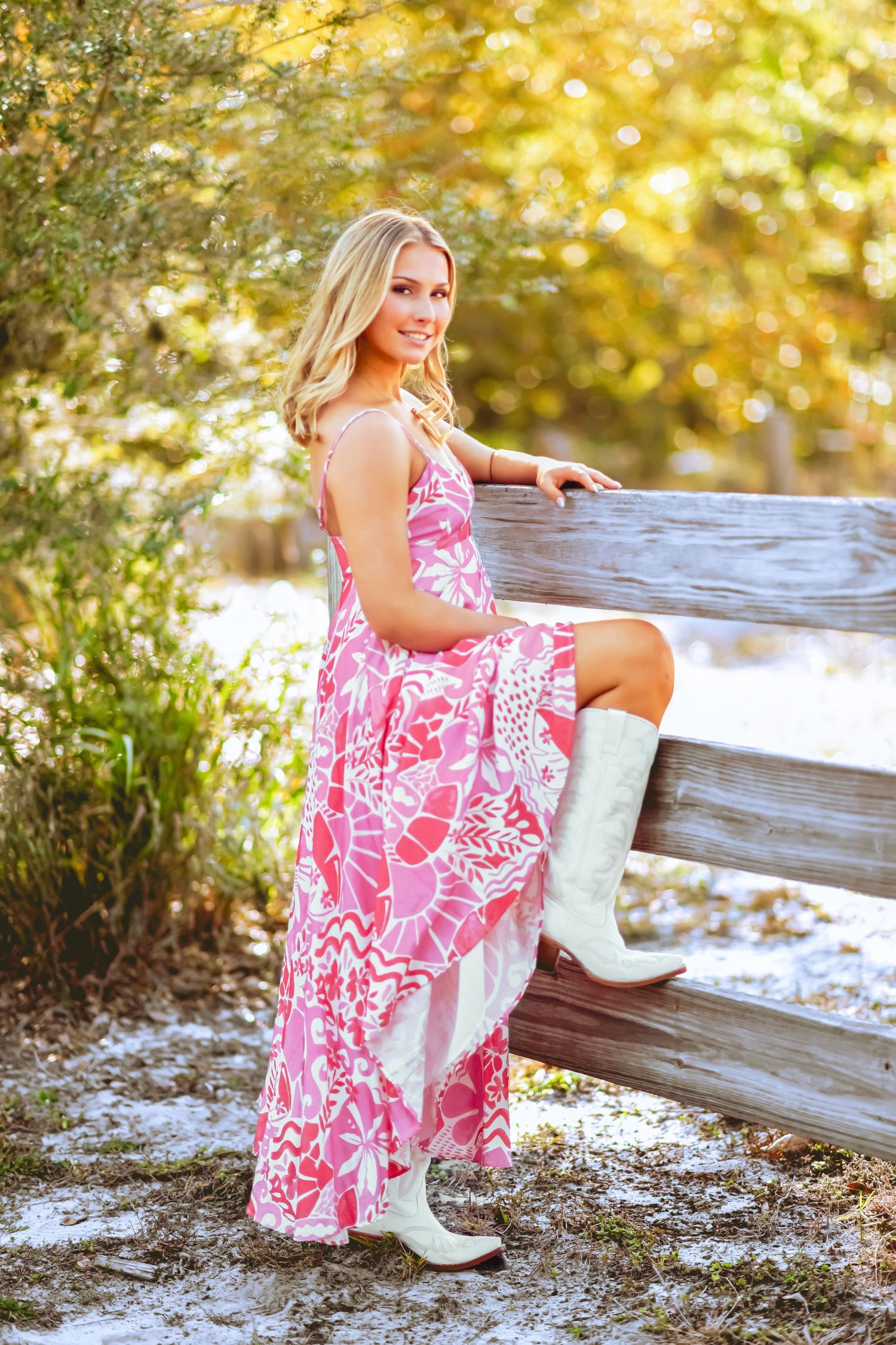 Blonde woman in pink floral maxi dress and white cowboy boots leaning on rustic wooden fence outdoors.