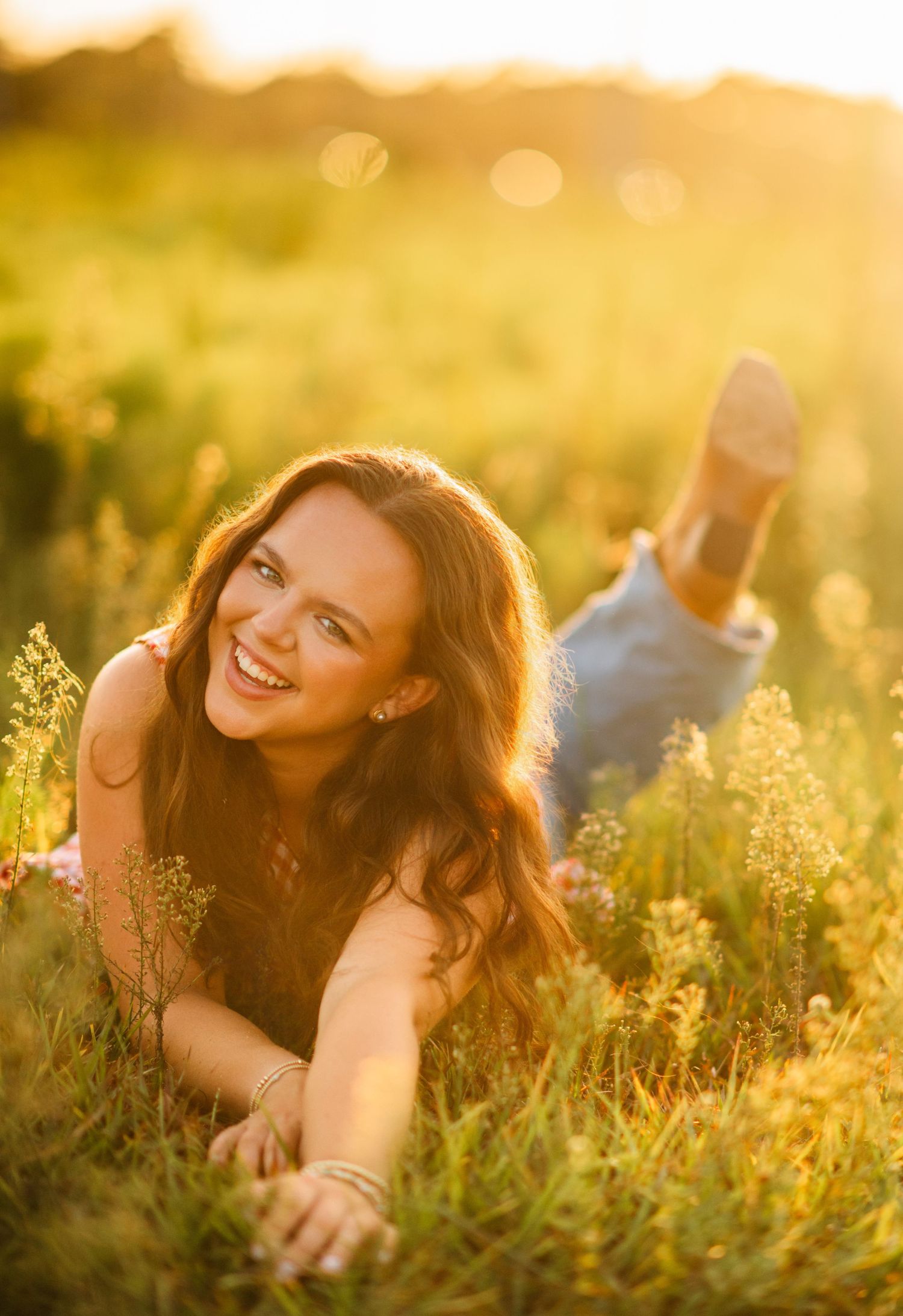Smiling young woman lying in golden sunlit meadow grass, wearing jeans, with long brown hair, enjoying a sunny day outdoors.