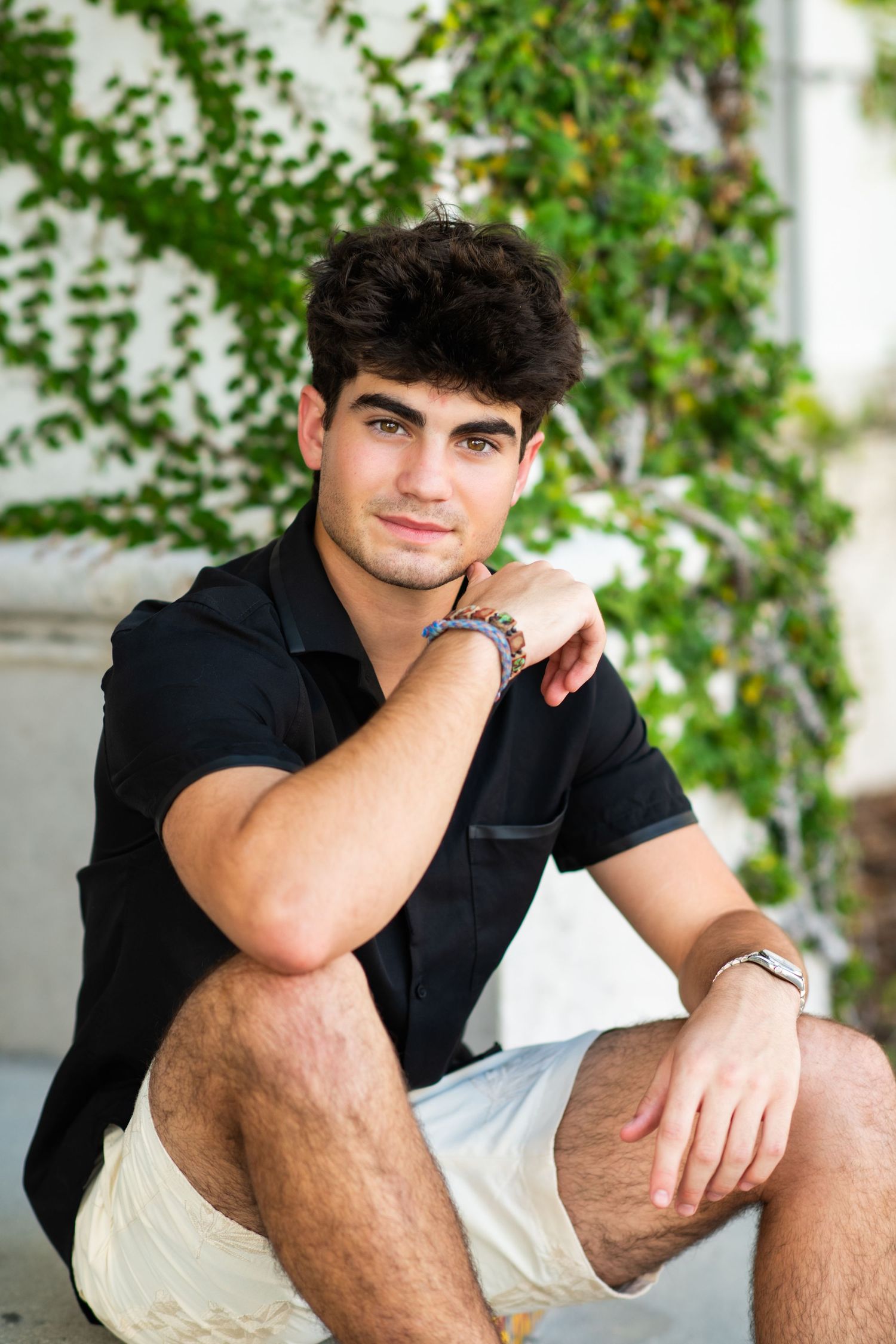 Young man in black shirt and white shorts sitting outdoors against ivy-covered wall, posing casually.