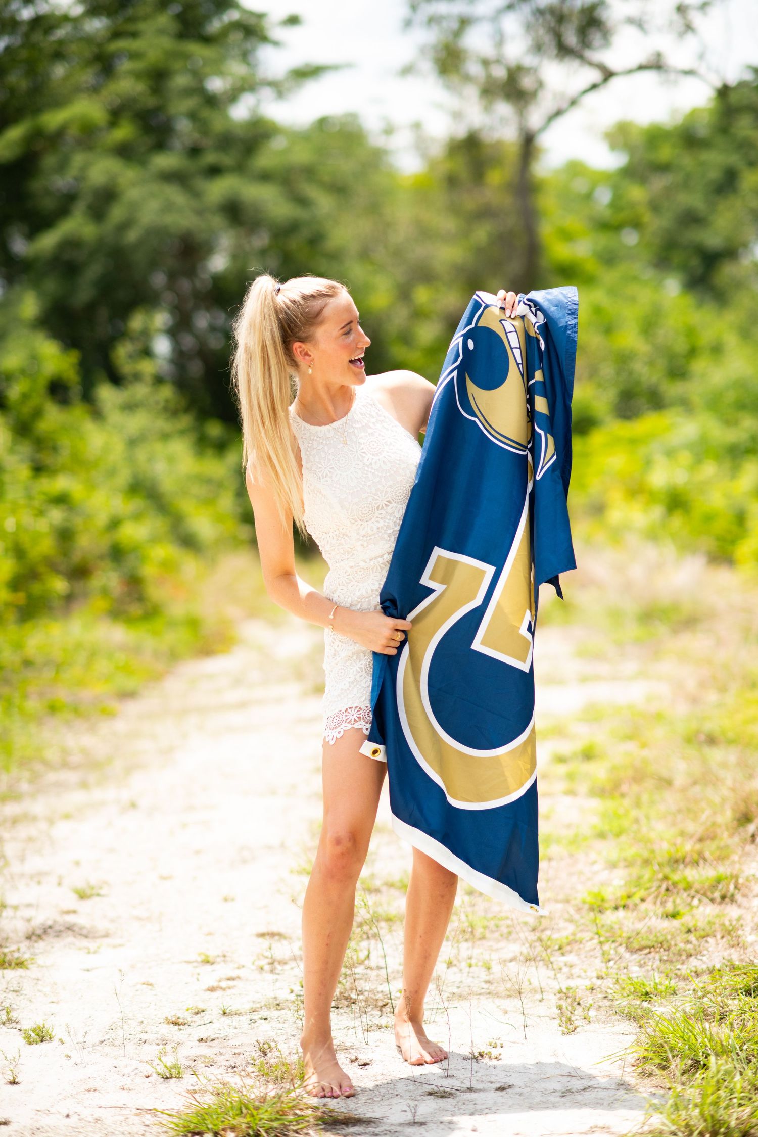 Blonde woman in white dress holding a blue and gold sports flag on a sandy path surrounded by greenery.