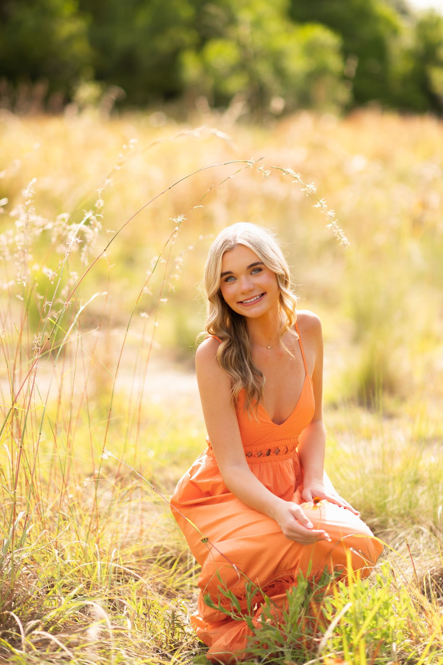 Young blonde woman in orange maxi dress sitting in golden field, smiling outdoors in warm sunlight.