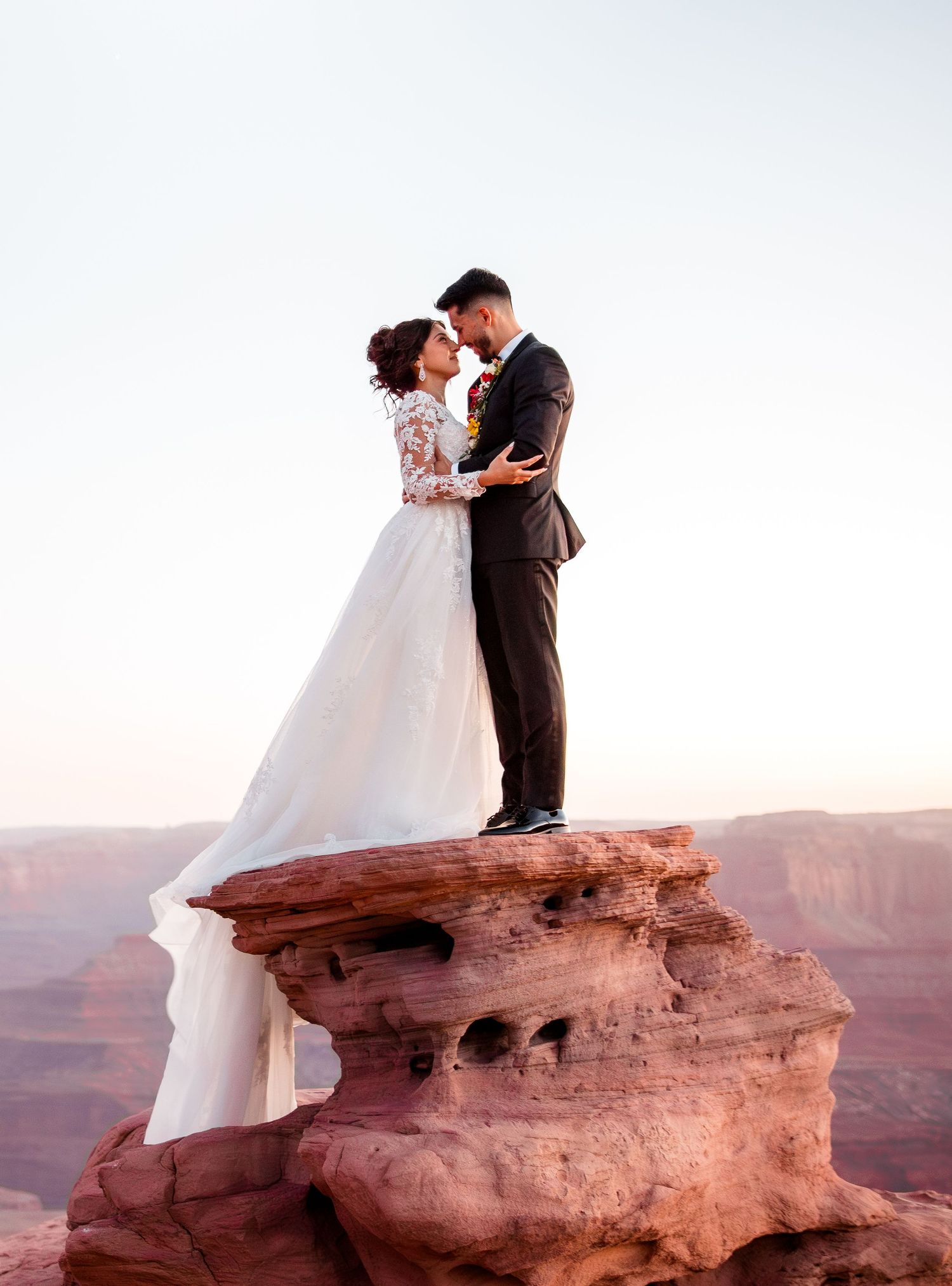 Couple in wedding attire embracing on red rock formation overlooking desert canyon.