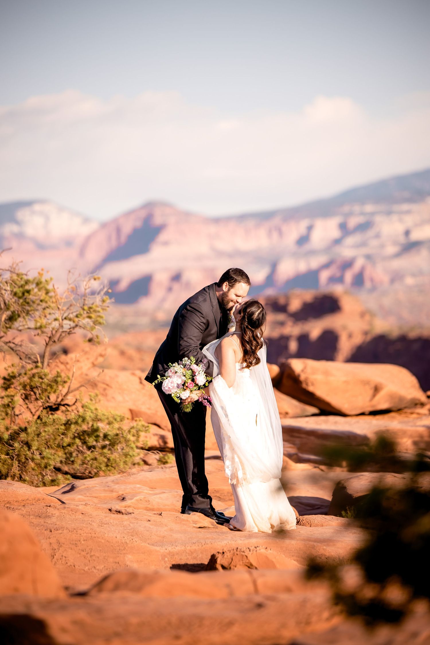 A wedding couple shares a romantic moment on red rock cliffs with mountains in the background.