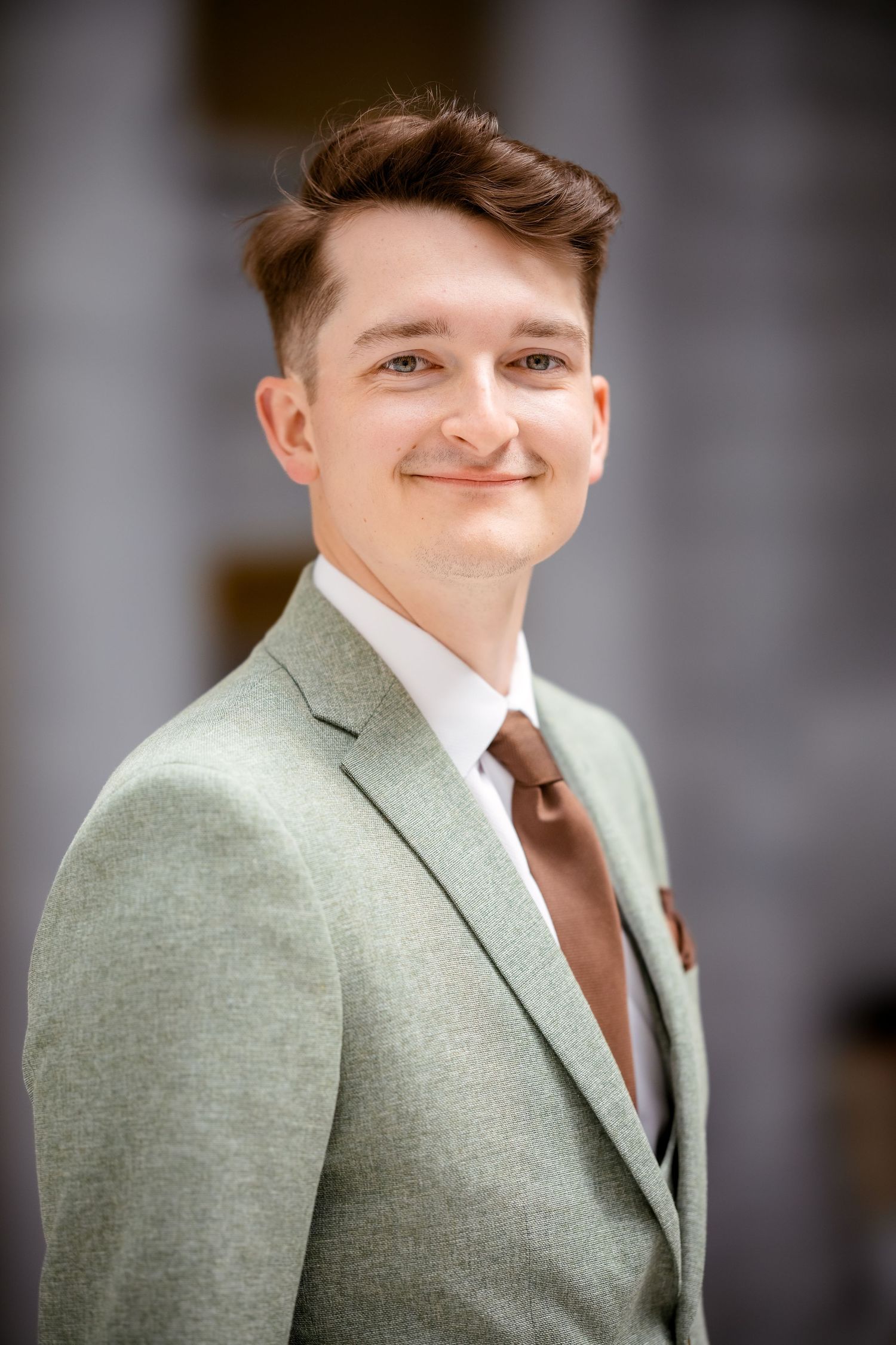 Groom wearing light sage green suit jacket with brown tie against blurred background.
