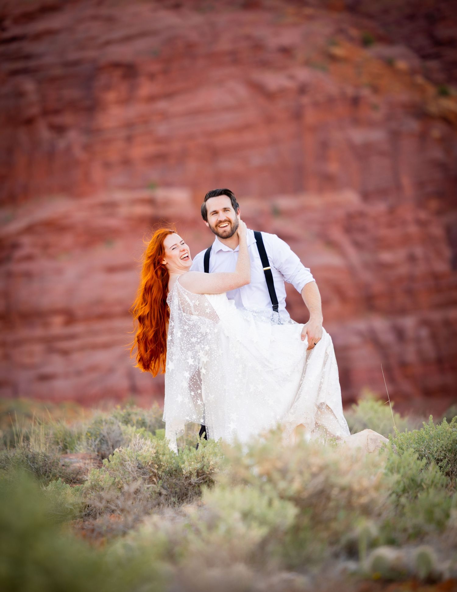 Newlyweds share intimate moments among desert brush with towering red cliffs in the background.
