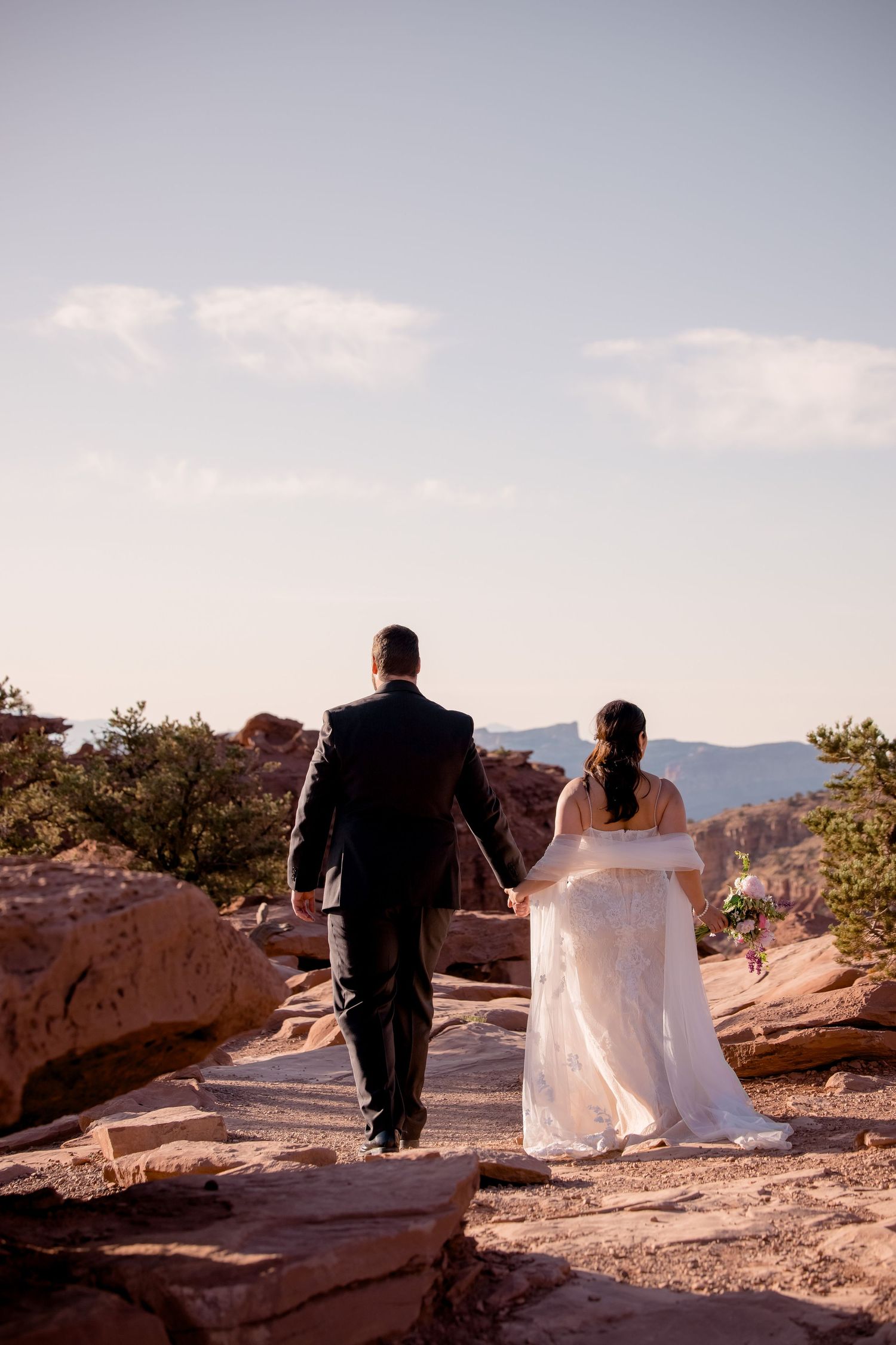 A couple walks hand in hand along rocky desert terrain at sunset.