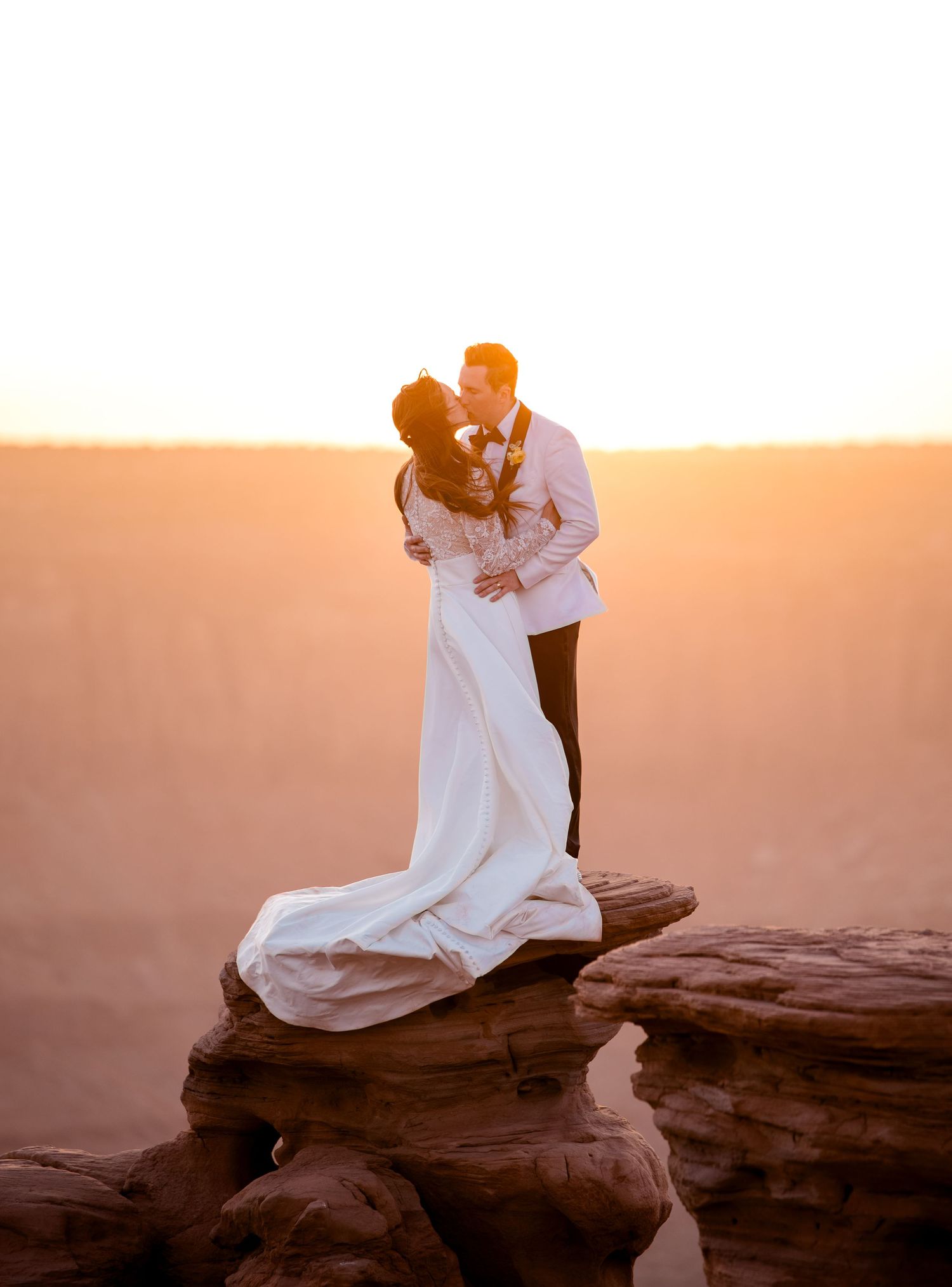 Romantic sunset embrace on red rock cliff with flowing white wedding dress.