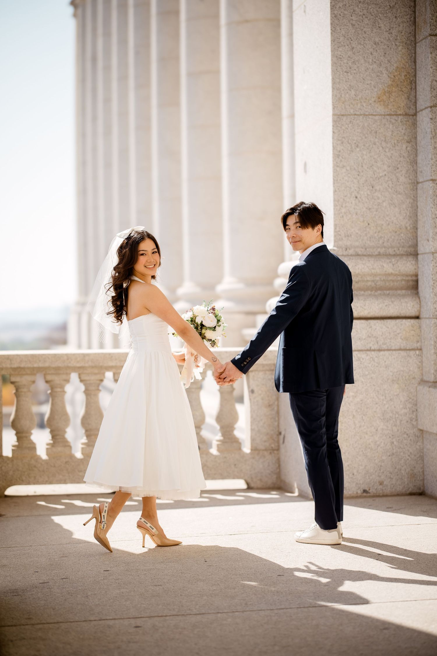 A playful wedding couple dances in a sunlit colonnade with classic architecture.