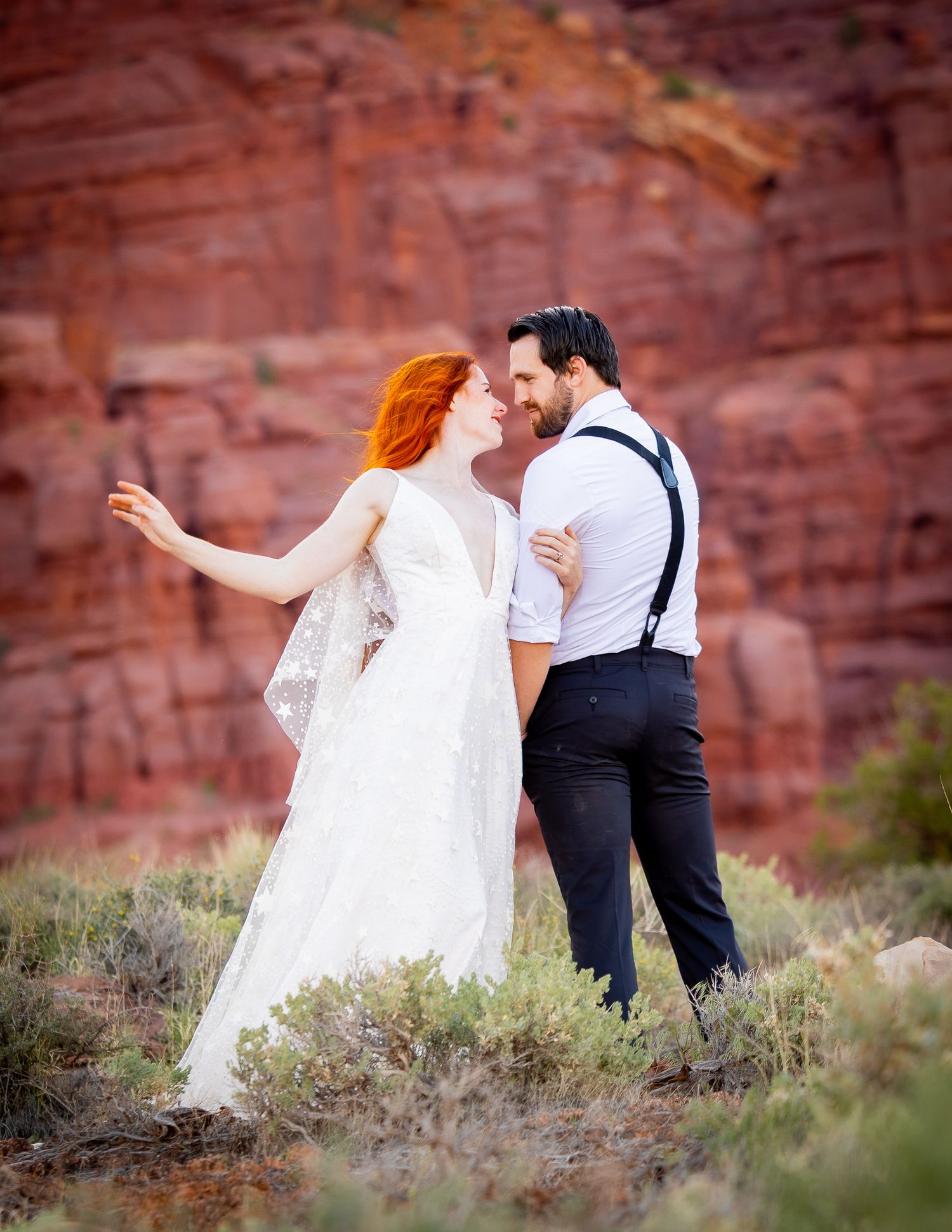 Wedding couple shares playful moments among desert vegetation with red canyon walls behind them.