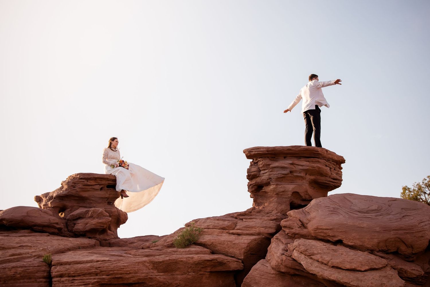 Wedding couple posing dramatically on separate red rock formations against bright sky.