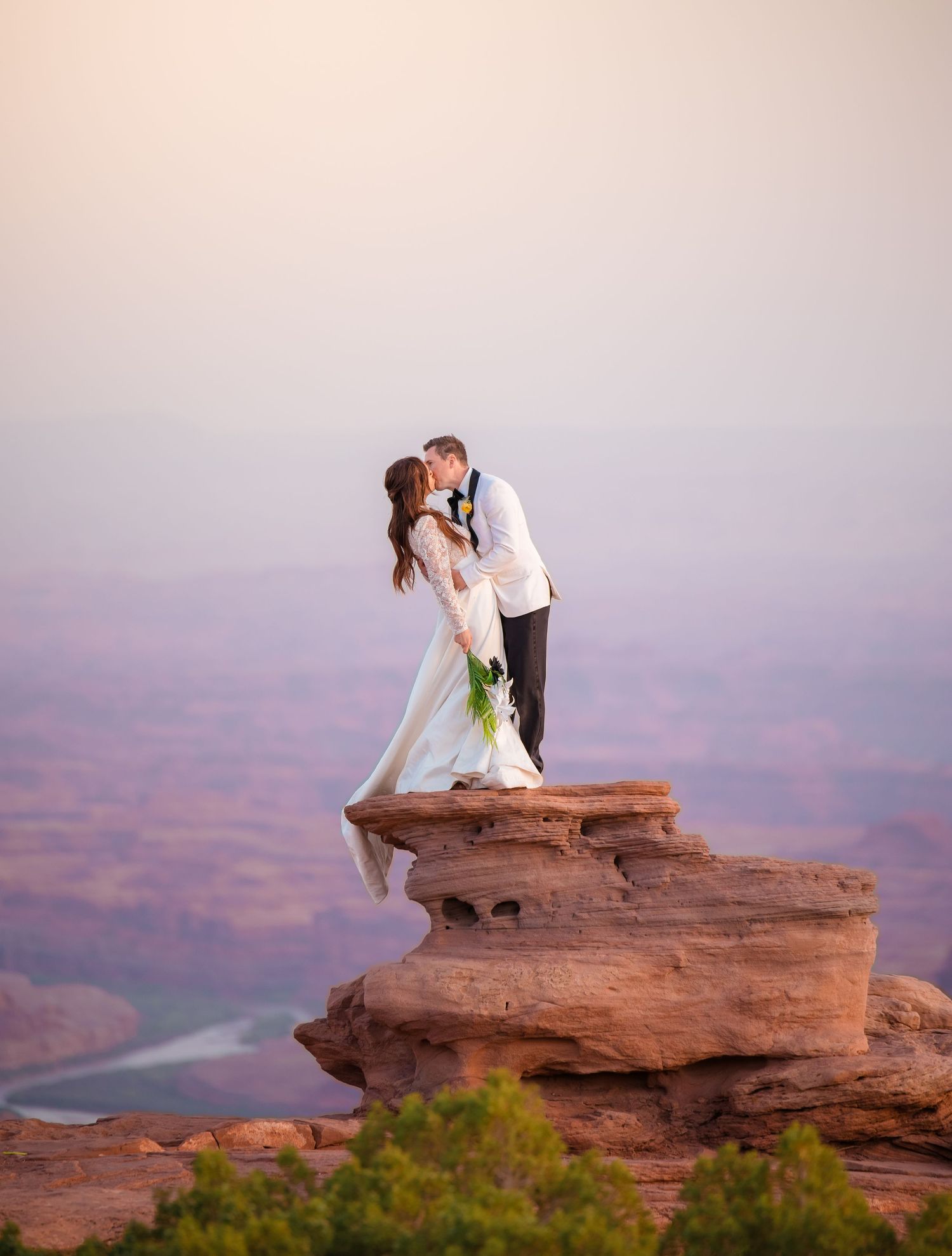 Wedding couple embraces on a dramatic red rock cliff edge overlooking a vast desert landscape.