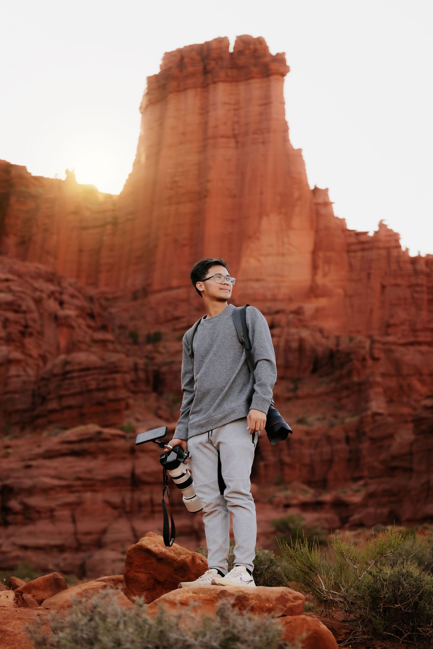 Person in grey clothing poses against towering red rock formations at sunset.