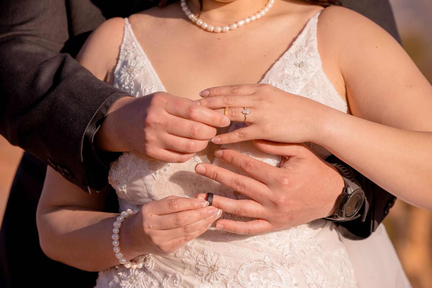 Close-up of hands with wedding rings and jewelry against a white dress.