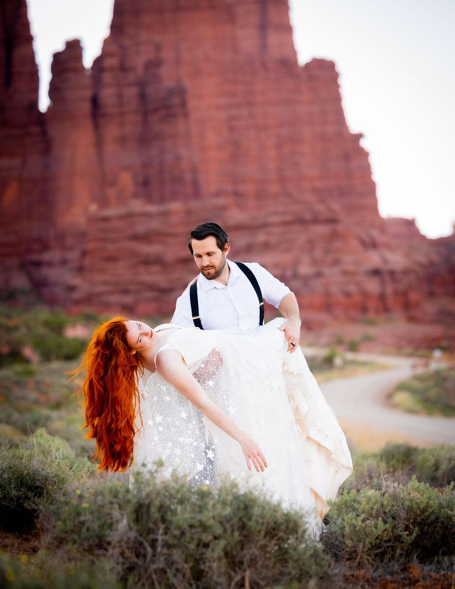A couple in wedding attire poses together against stunning red rock formations in a desert landscape.