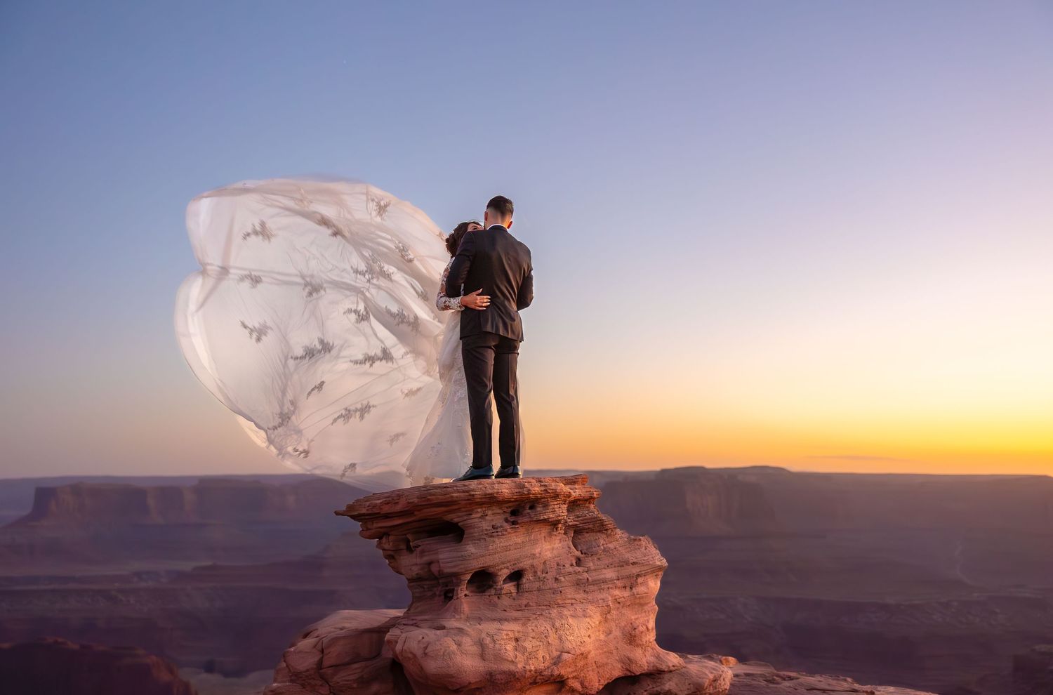 Figure standing on red rock formation with flowing white fabric during desert sunset.