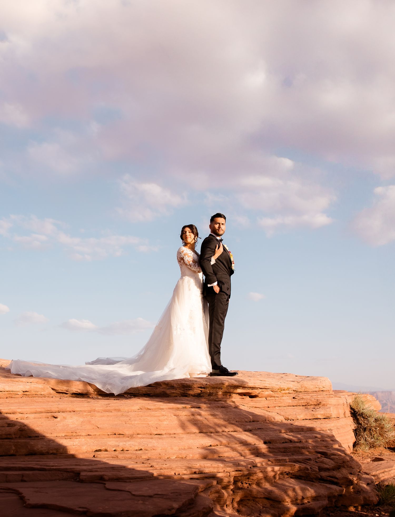 A couple in wedding attire poses dramatically on red rock formations in the desert at sunset.