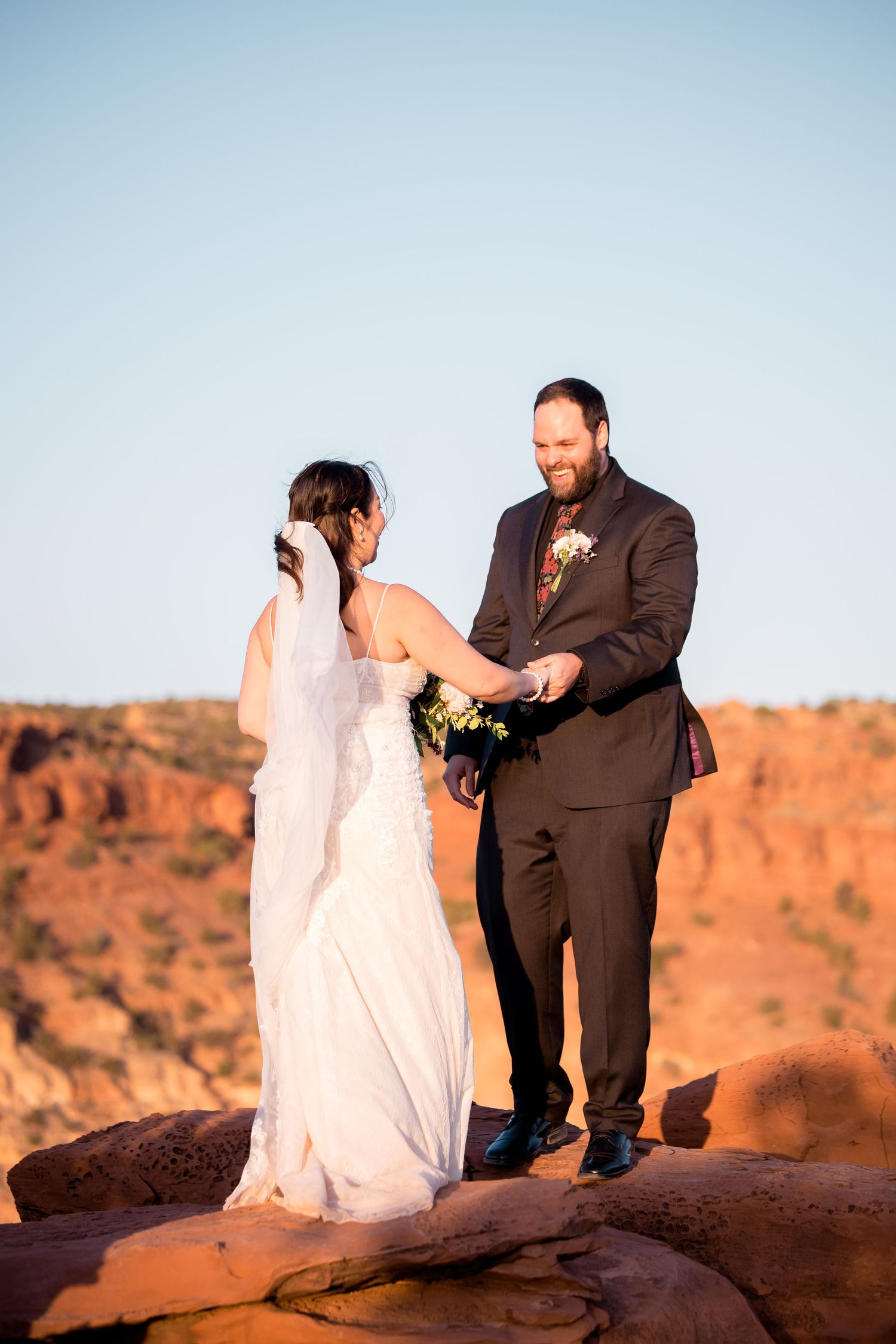 A wedding couple poses on red rock formations during sunset in a desert landscape.