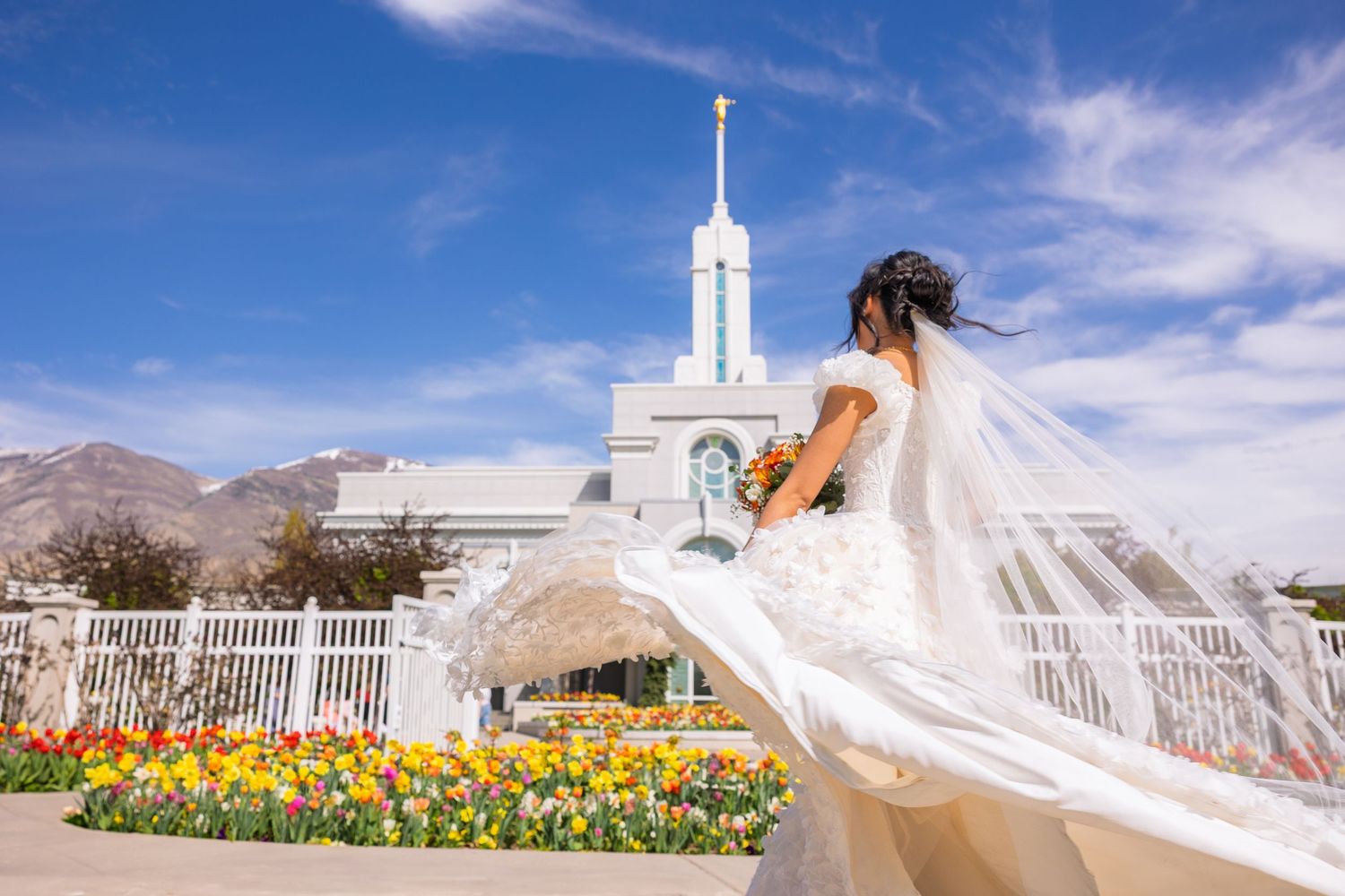 A flowing white dress catches the wind against a white temple and yellow tulip garden backdrop.