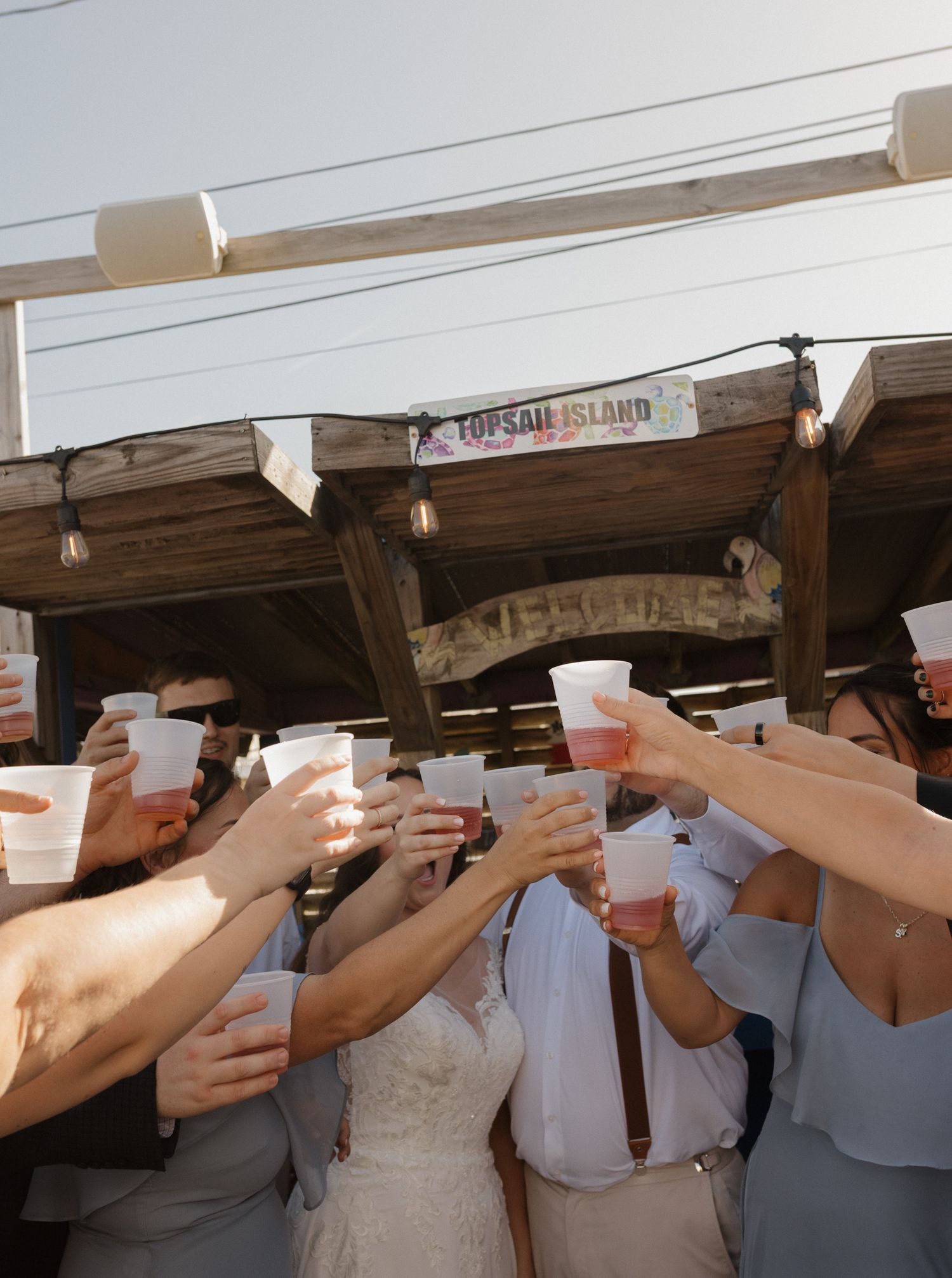 Wedding guests toasting with drinks under wooden beams and string lights.