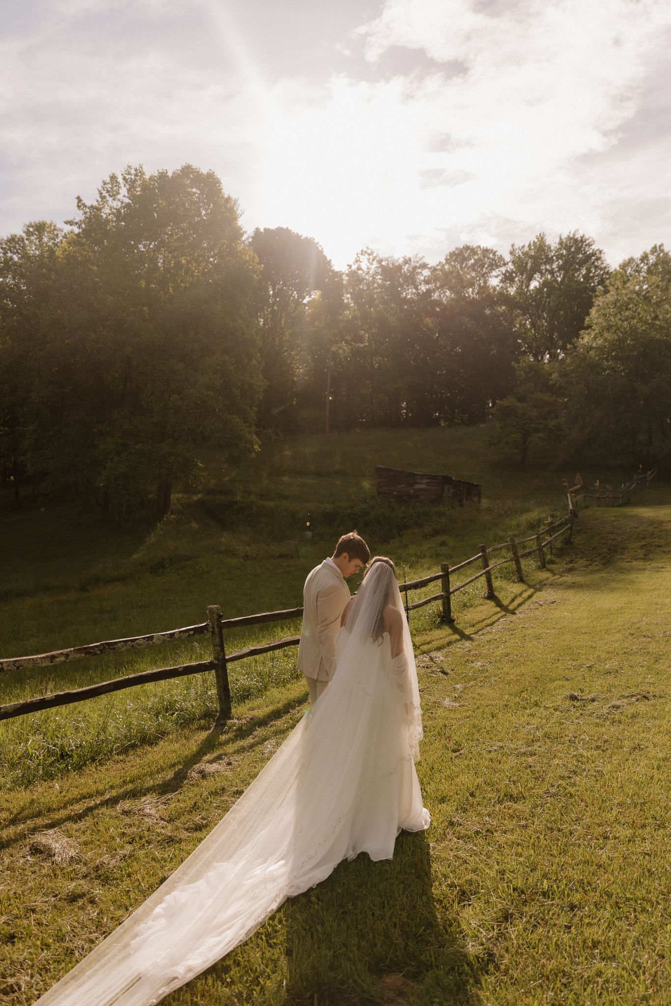 Silhouetted wedding couple against sunset with dramatic veil on rolling hillside.