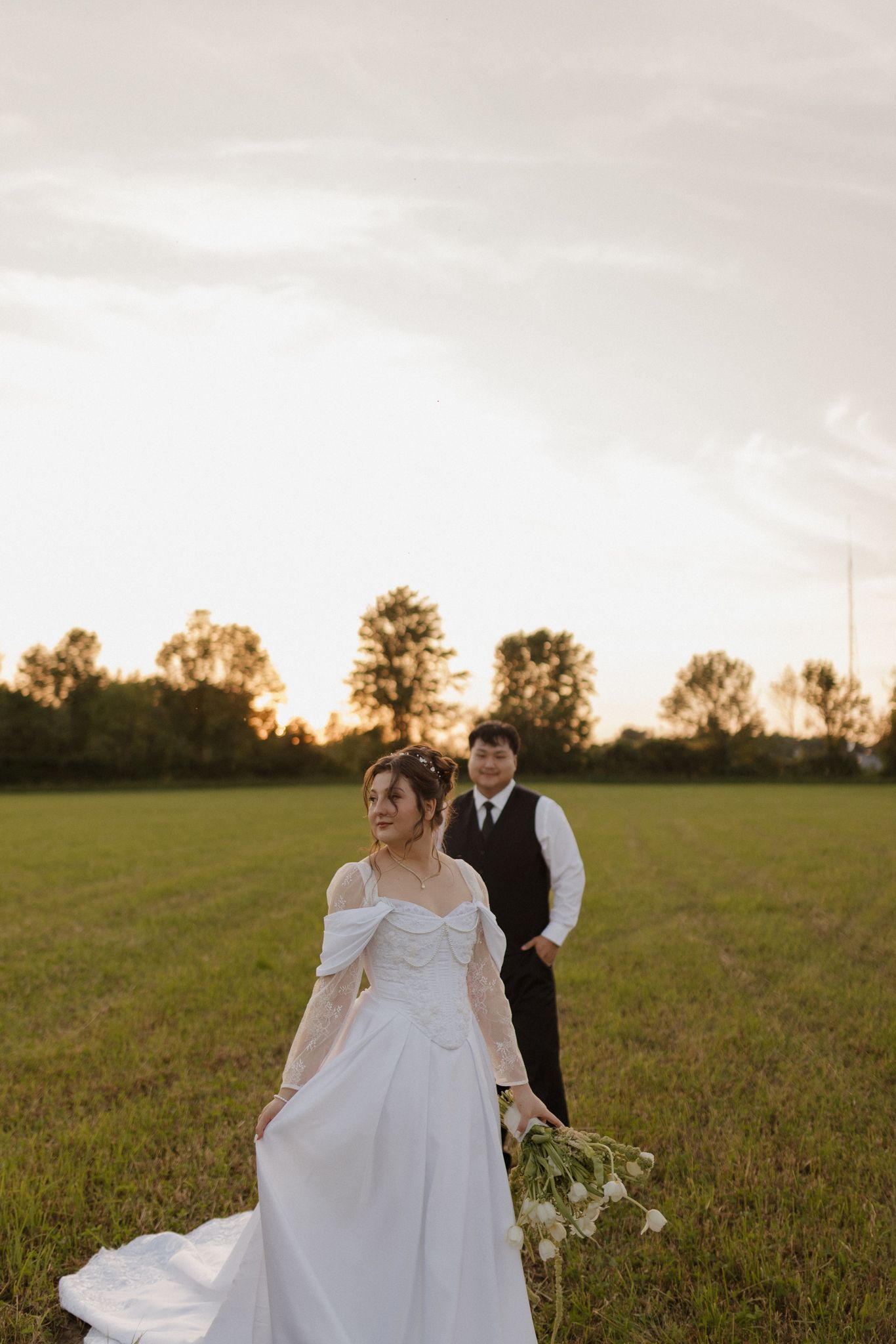 A bride in a flowing white dress poses in a field during golden hour while a groom stands behind her.