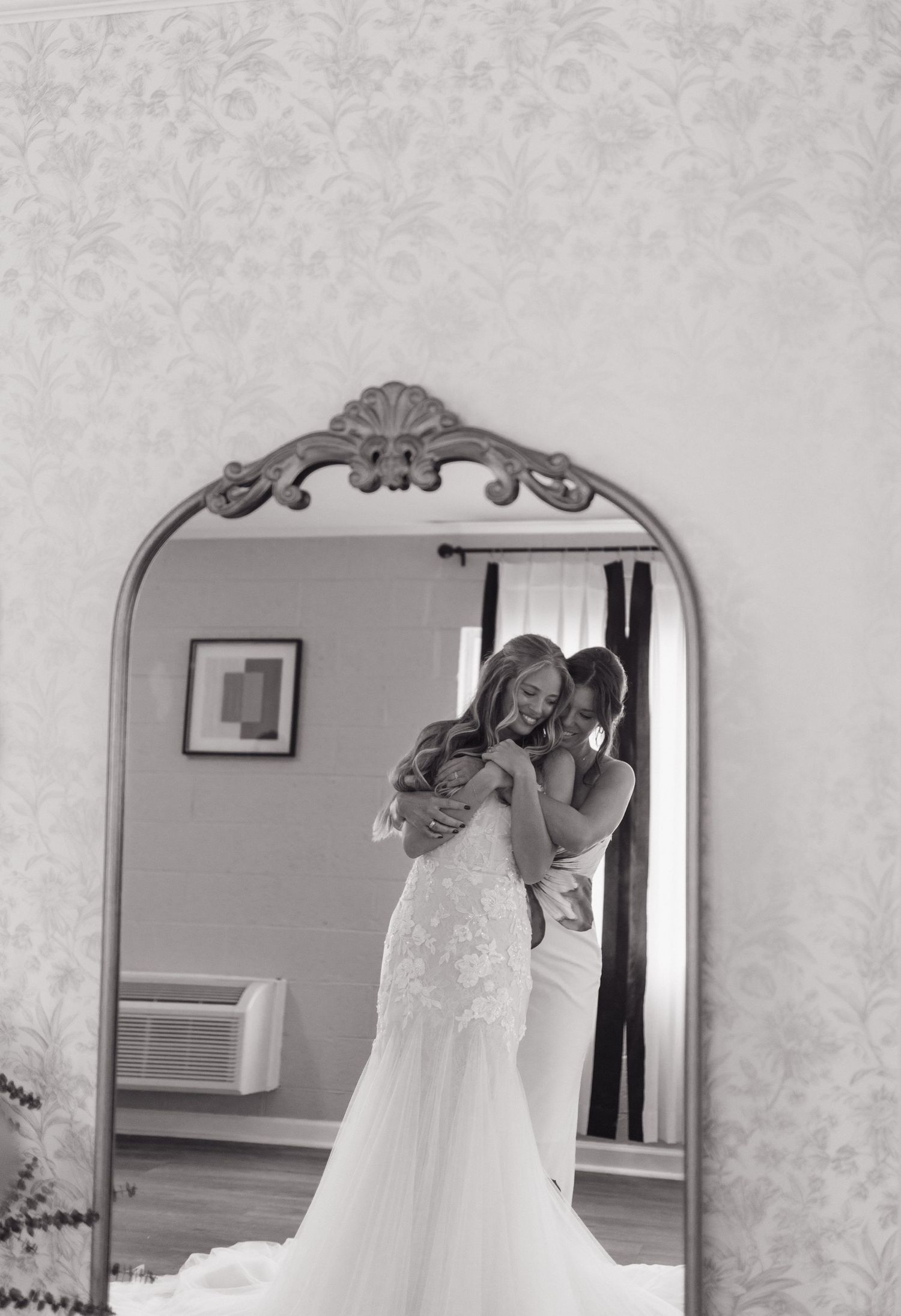 Black and white reflection in ornate mirror shows a romantic moment in a wedding dress fitting room.