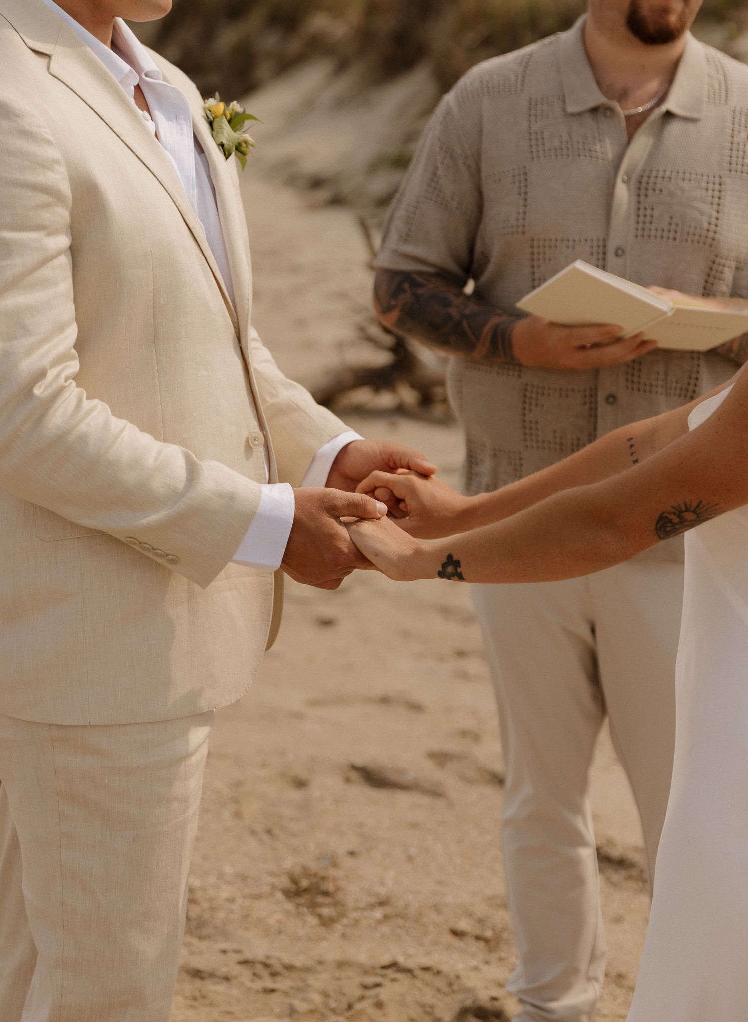 Close-up detail of groomsmen wearing beige suits and watching a ring exchange during an outdoor beach wedding ceremony.
