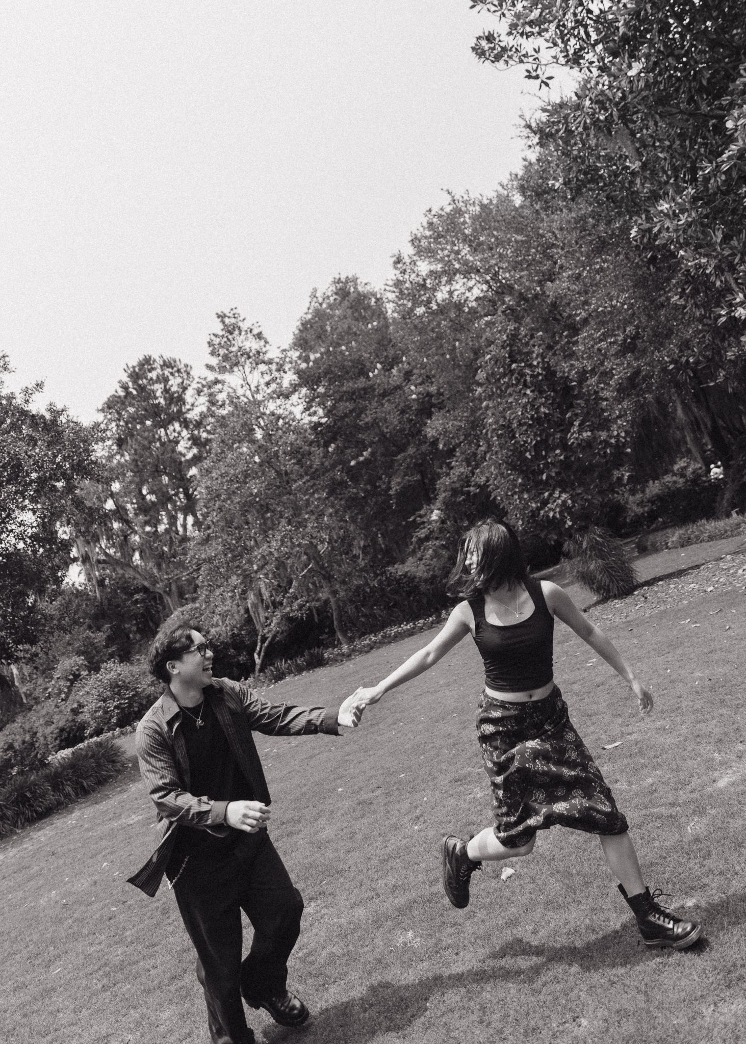 Playful couple holding hands while dancing on a grassy hillside in black and white.