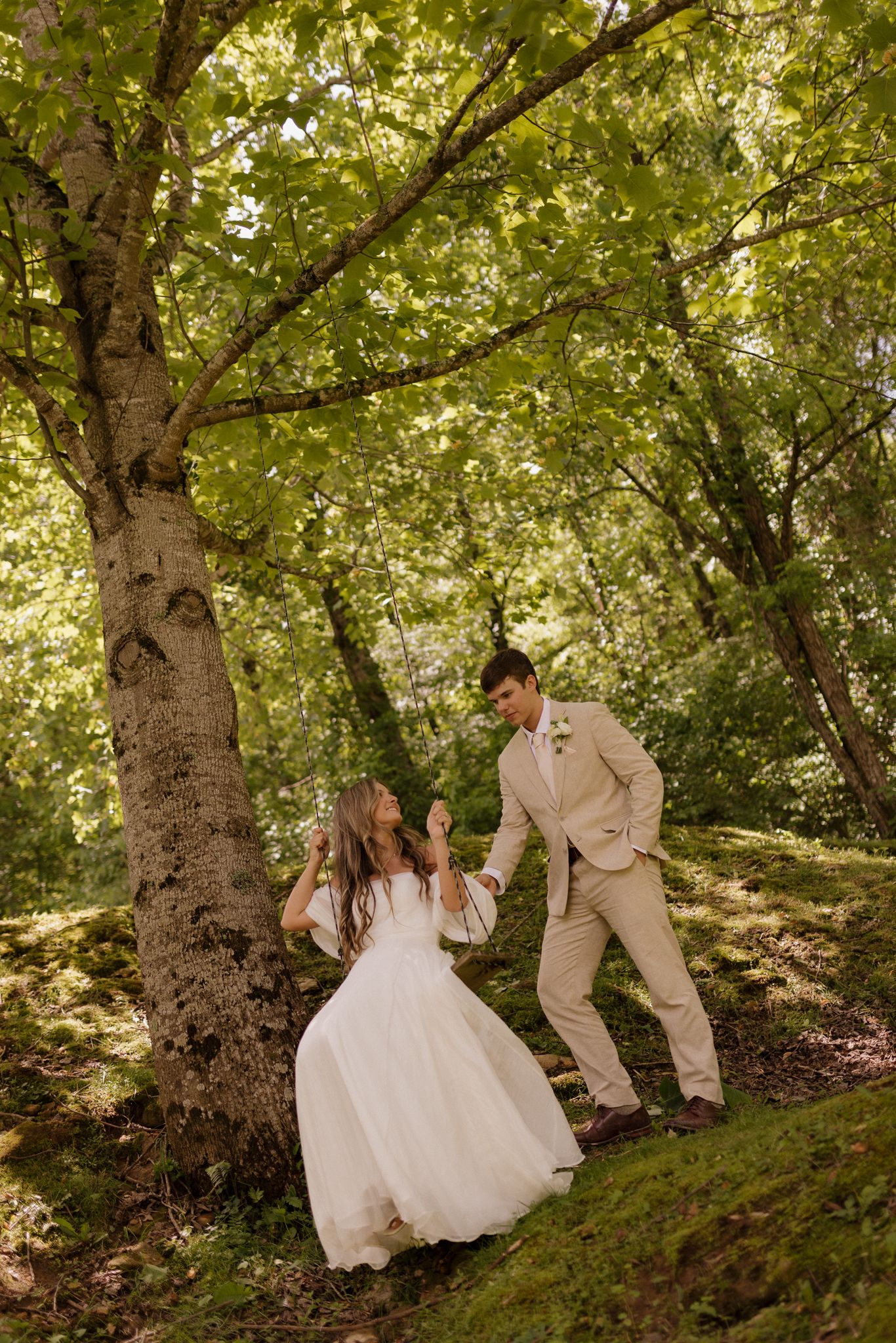 A couple in wedding attire poses playfully on a mossy hillside beneath large trees.
