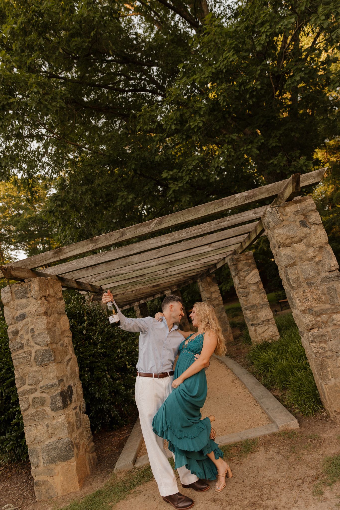 Dancing couple in formal attire beneath a rustic stone pergola at sunset.