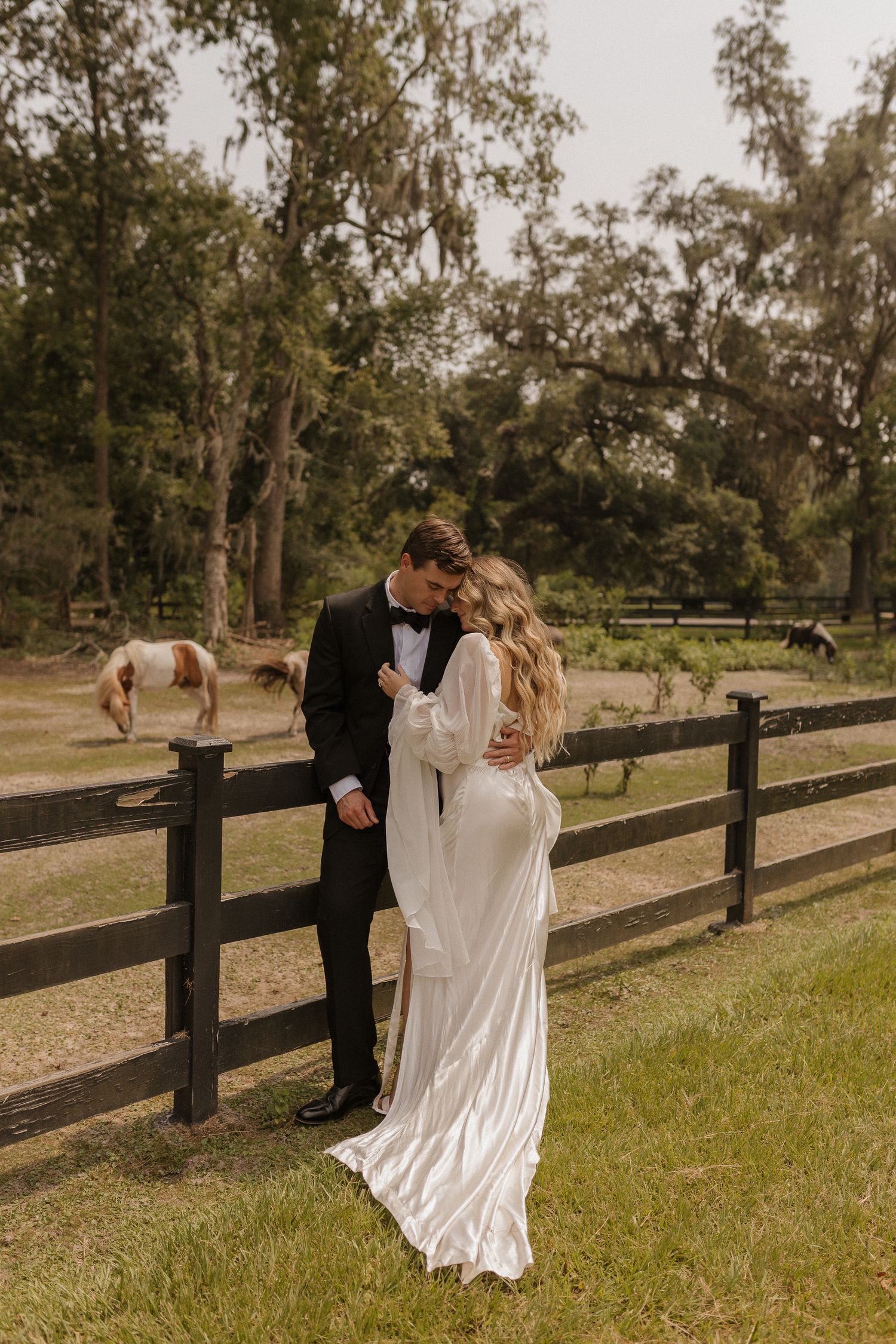 A couple in wedding attire embraces near a wooden fence at a rustic farm setting with a horse visible in the background.